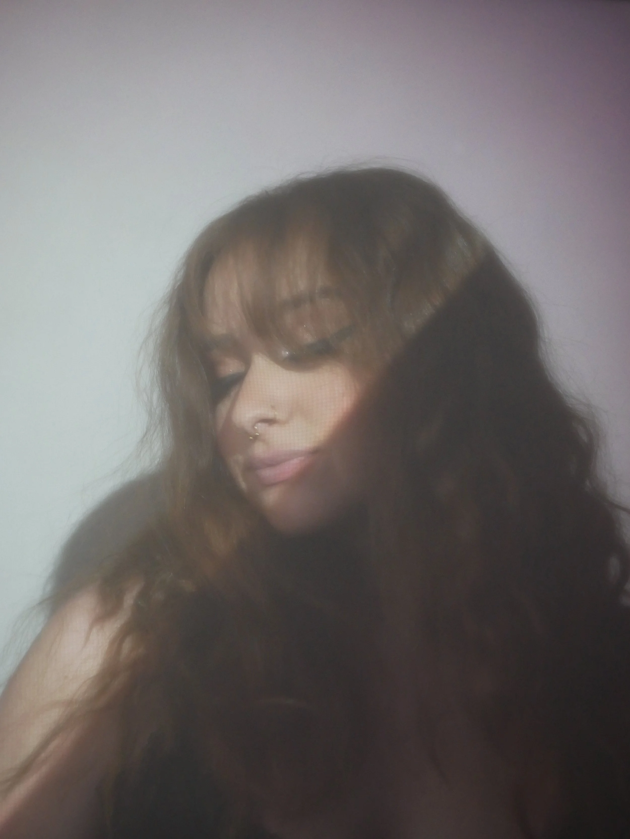 Portrait of a woman with long dark curly hair, wearing makeup and a septum piercing, with her eyes closed and slight smile, against a plain light background.