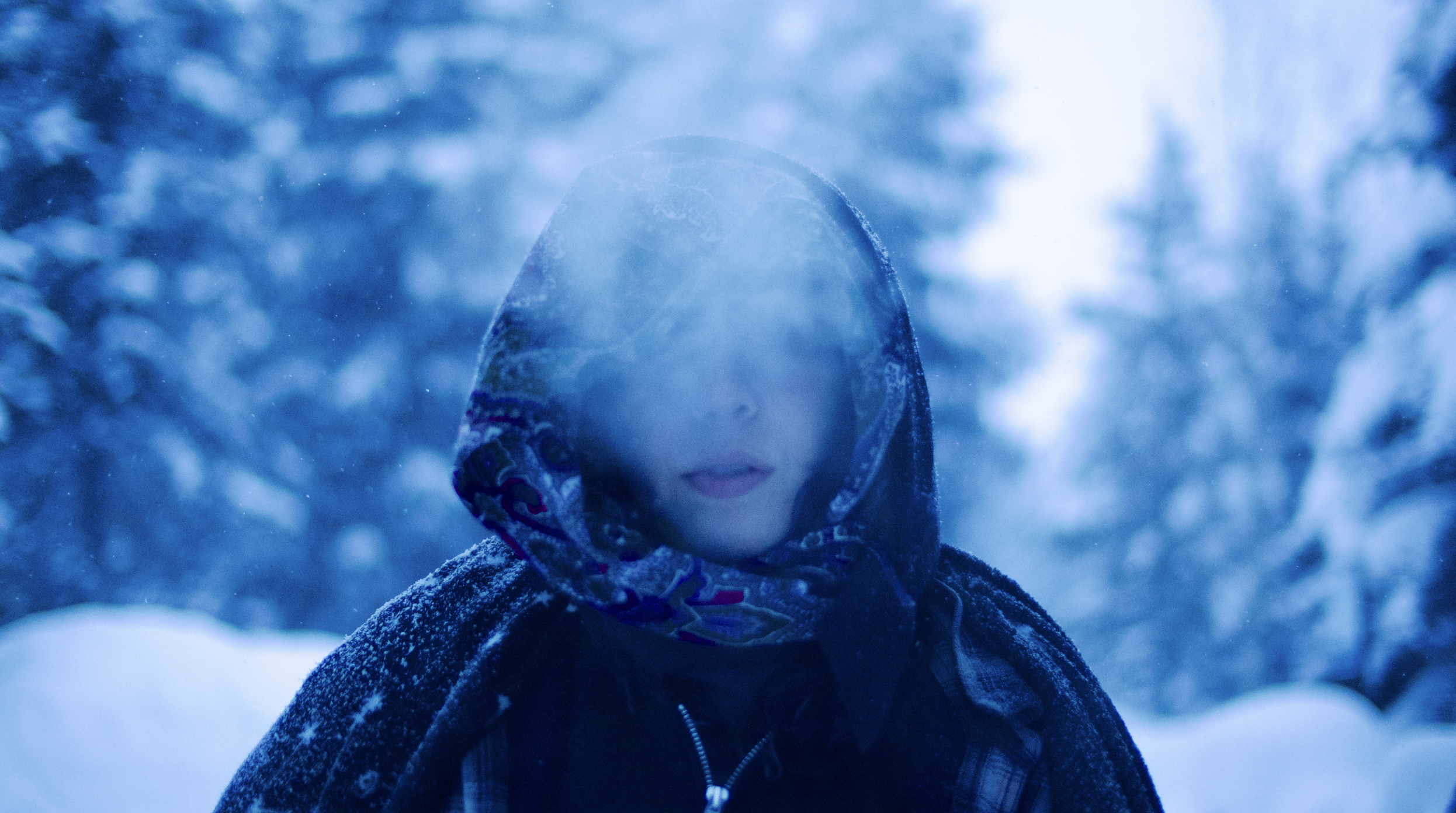 Person wearing a hooded jacket and scarf standing outdoors in a snowy forest, with a blurred background of snow-covered trees.