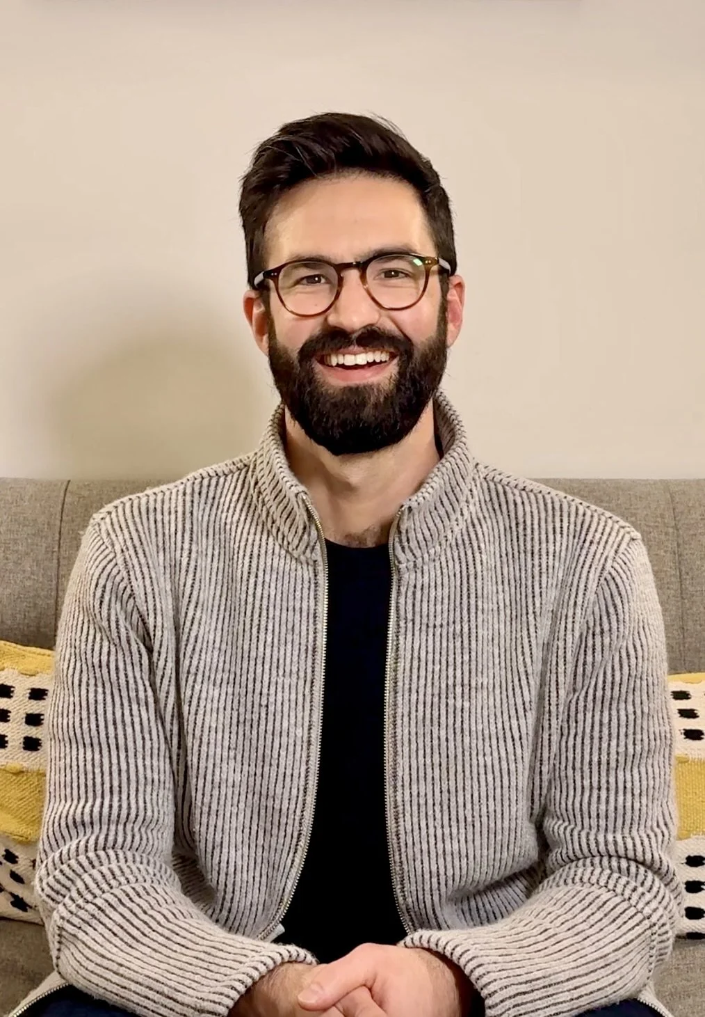 A smiling man with glasses and a beard, wearing a gray zip-up sweater, sitting on a beige couch with yellow and black patterned pillows against a plain wall.