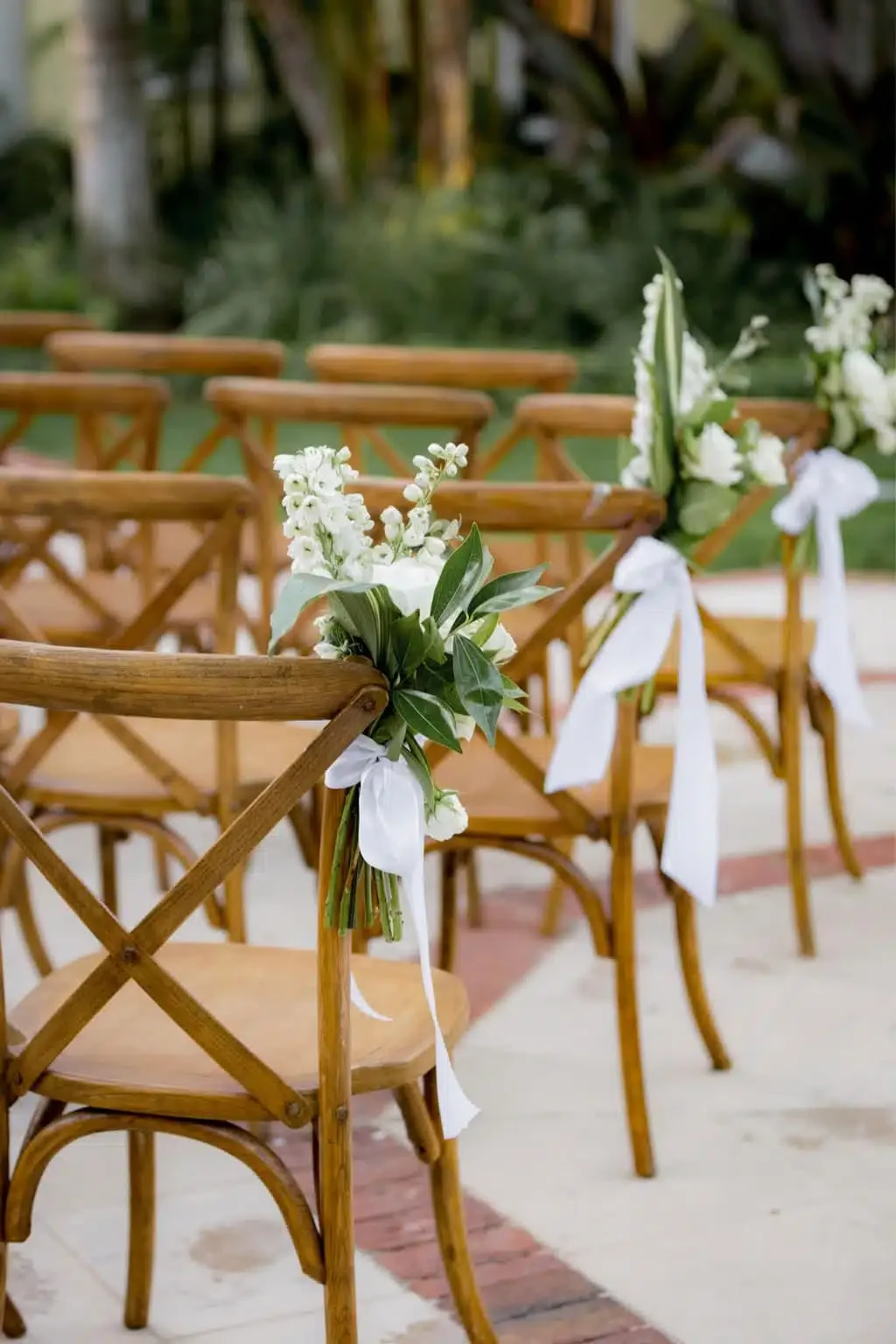 Wooden chairs decorated with white flowers and green leaves tied with white ribbons, arranged for a wedding ceremony outdoors.