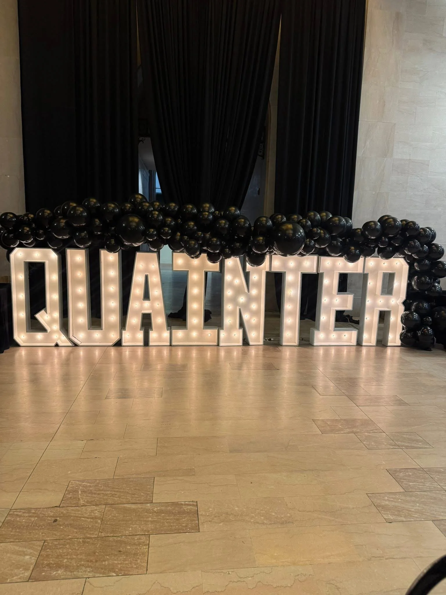 Large illuminated sign spelling 'QUANTER' with black balloons on top, set against dark curtains in an indoor space.