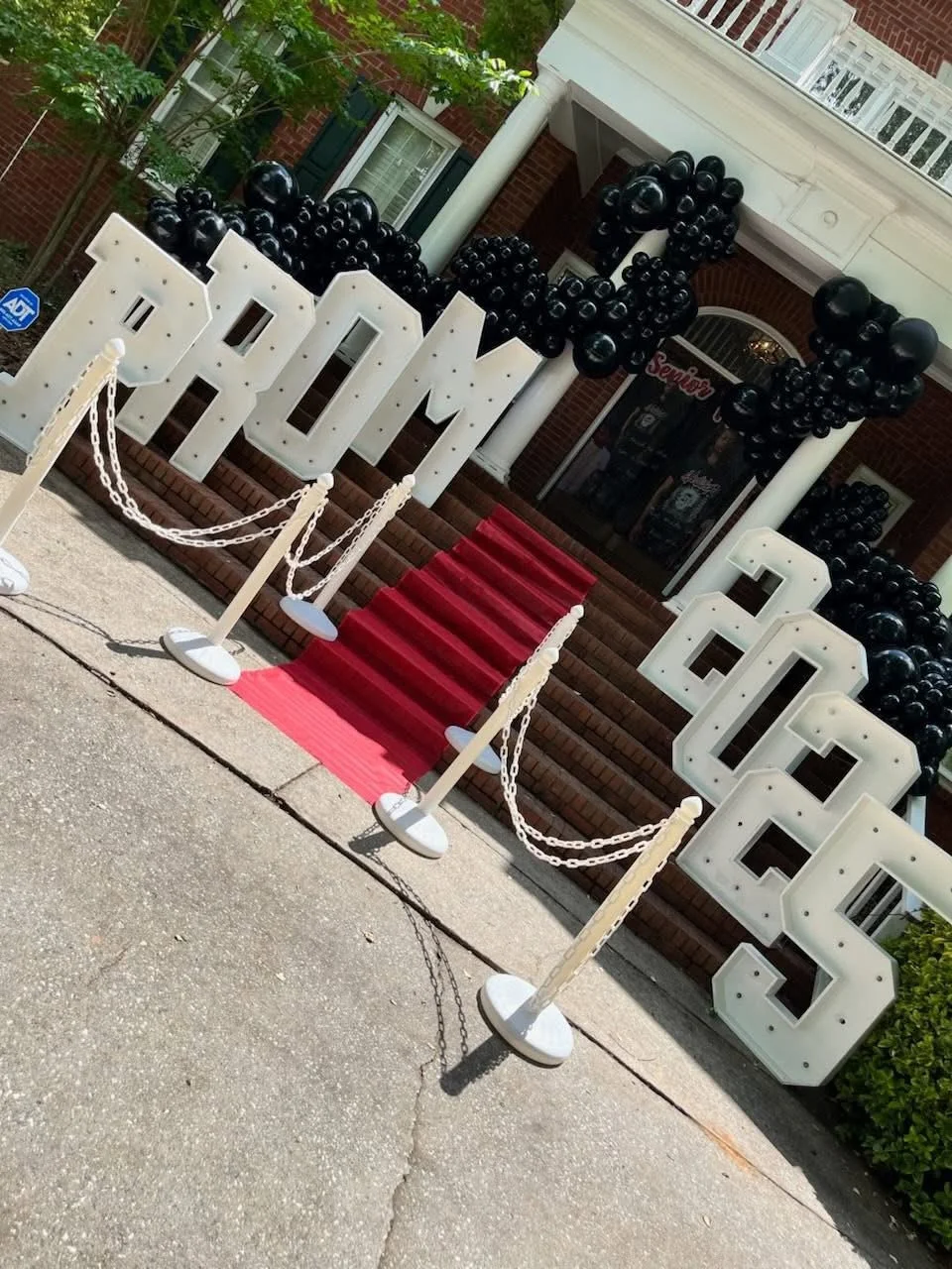 Decorative entrance setup with large white marquee letters spelling 'PROM' on steps, black balloon clusters, a red carpeted staircase, and white stanchions with chains