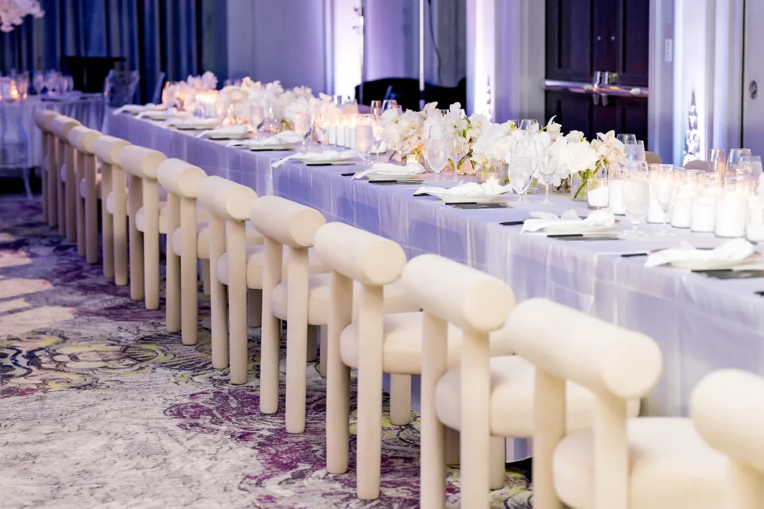 Banquet table set for an event with white tablecloths, floral centerpieces, candles, glassware, and place settings, with white chairs along one side.