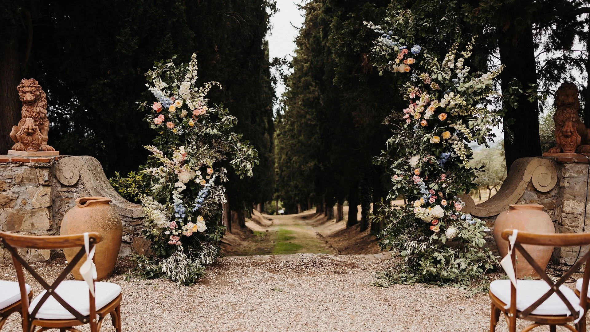 Outdoor wedding arch decorated with flowers, flanked by large terra cotta vases and wooden chairs, set against a path lined with tall trees.