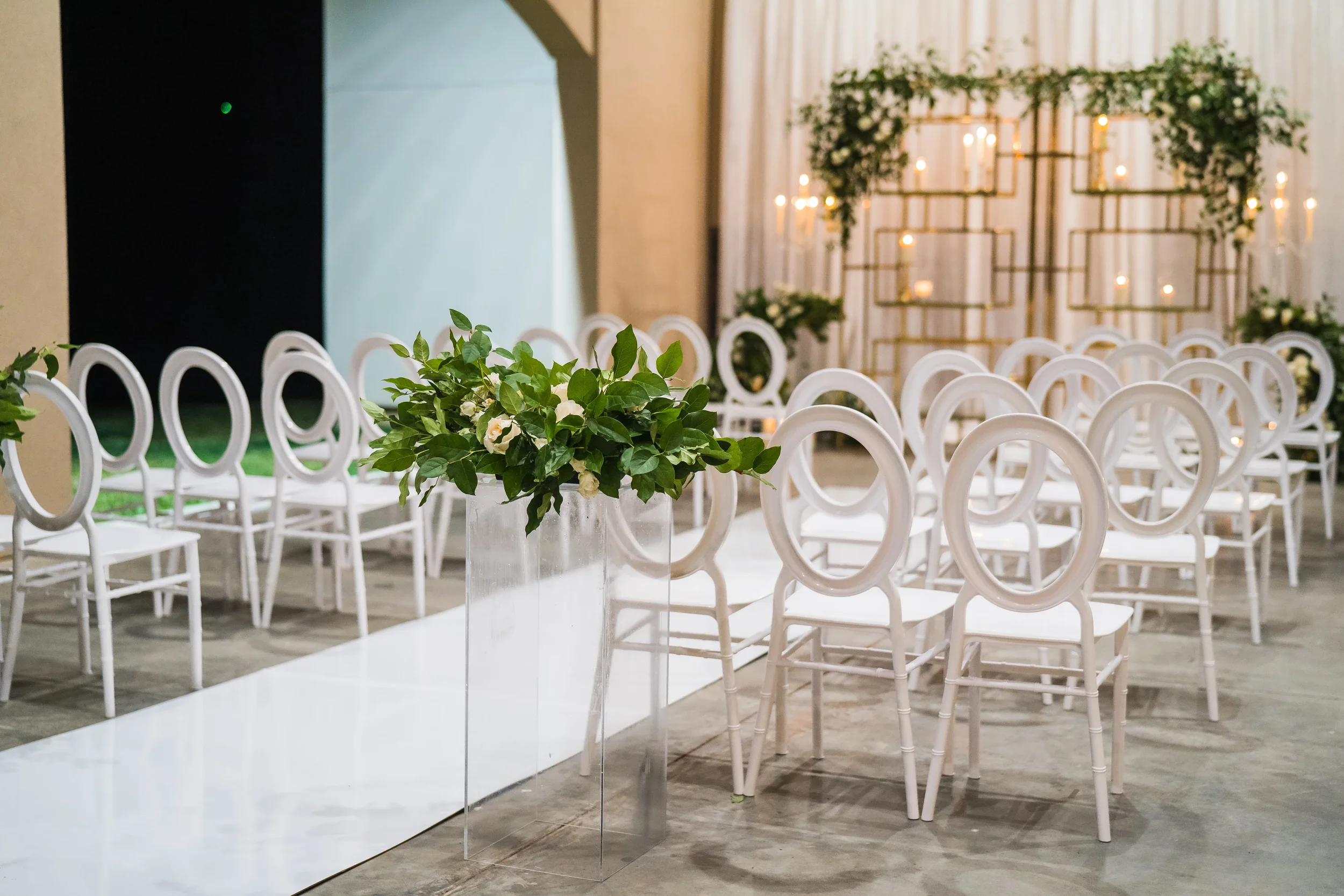 Wedding ceremony setup with white chairs, a flower arrangement in a clear vase, and a backdrop with candles and greenery.