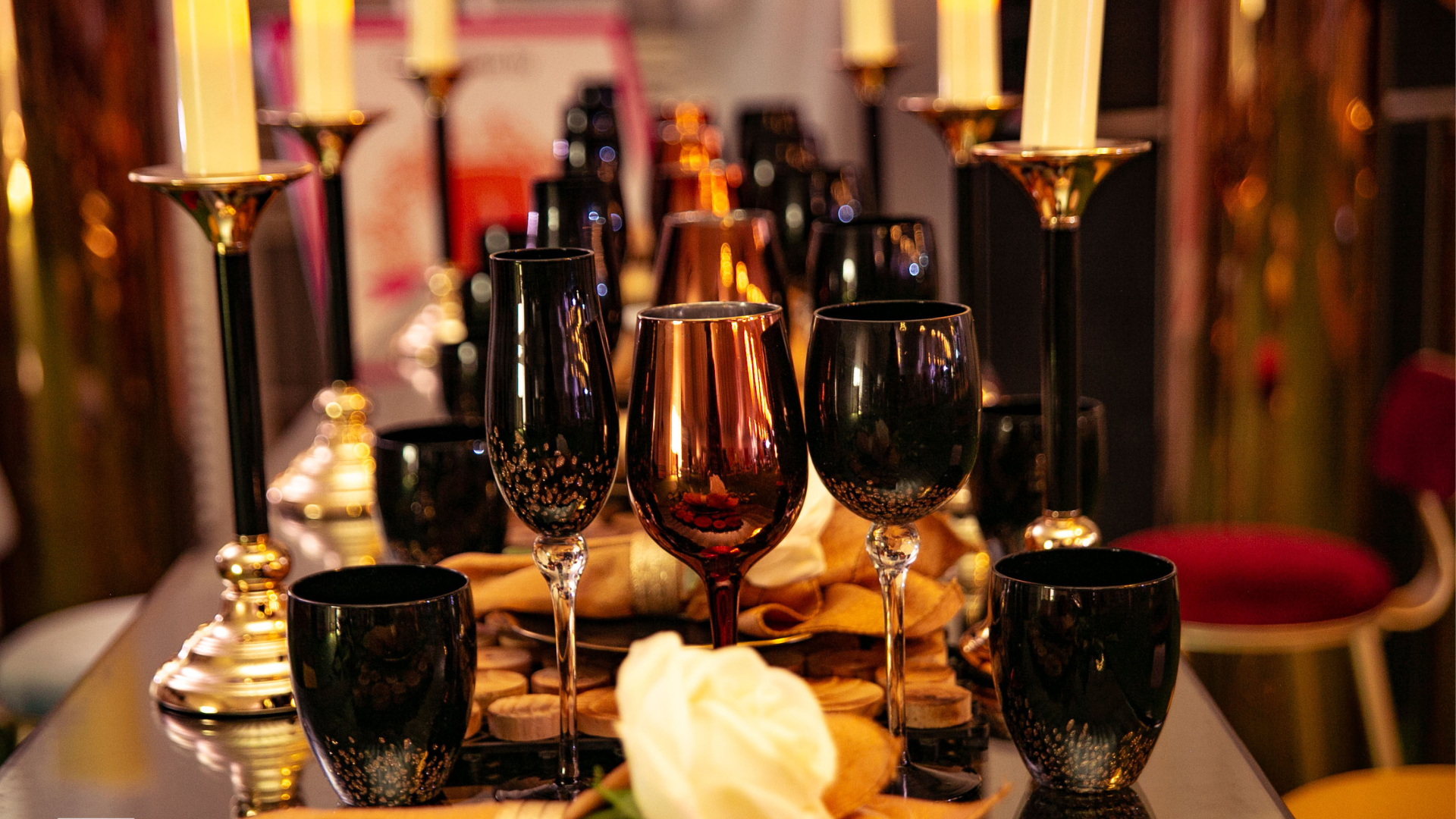 Elegant table setting with black and metallic glassware, tall white candles in gold holders, and a single white rose on a decorated table.