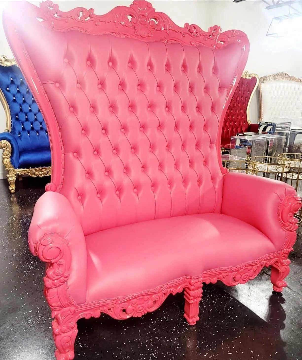 A pink Victorian-style high-back sofa with ornate wood carvings, tufted upholstery, and curved armrests, positioned in a furniture store among other colorful chairs.