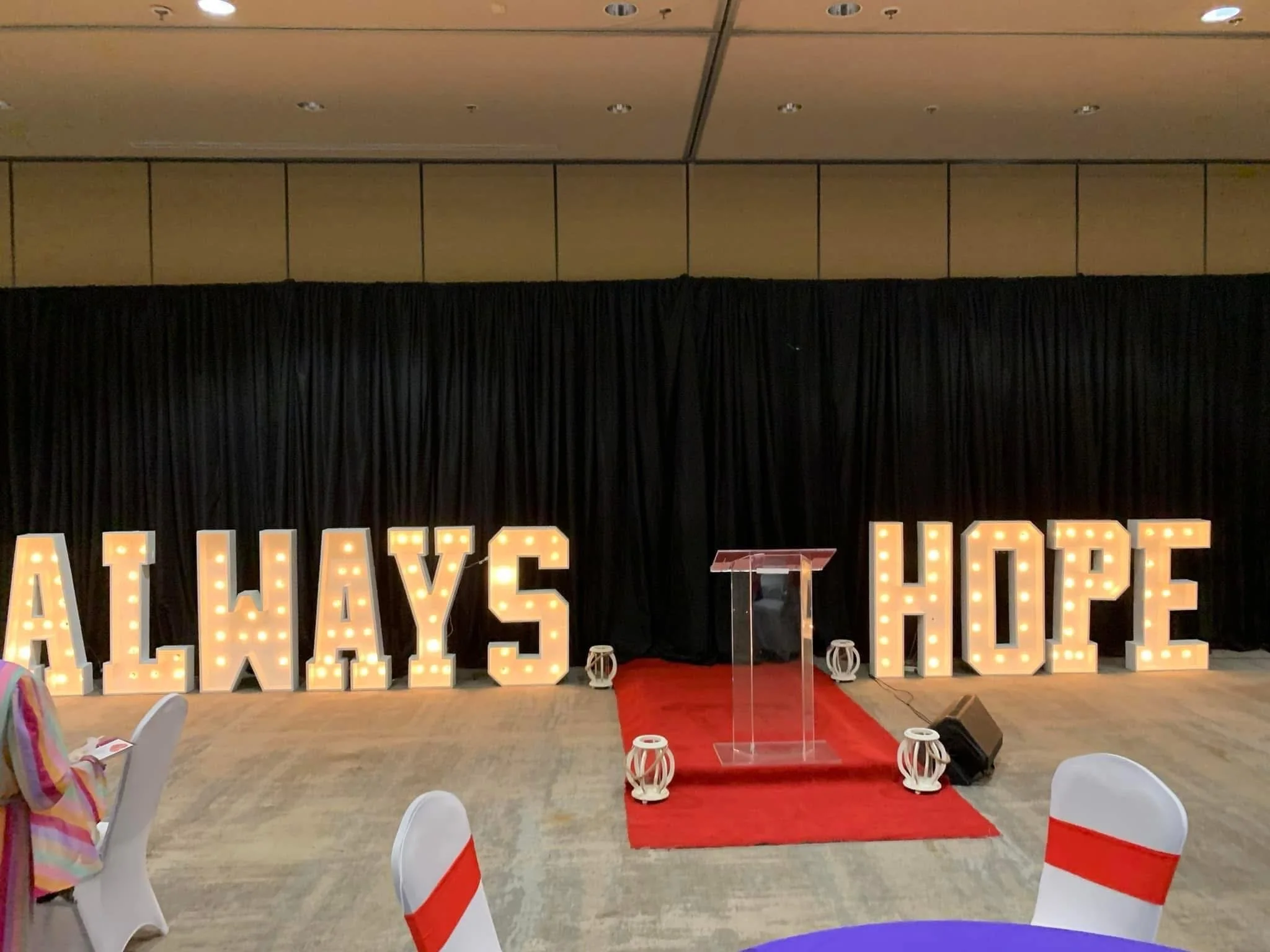 Decorative sign spelling out 'ALWAYS HOPE' with illuminated marquee letters, set on a stage with black curtain backdrop, a red carpet, and a clear podium.