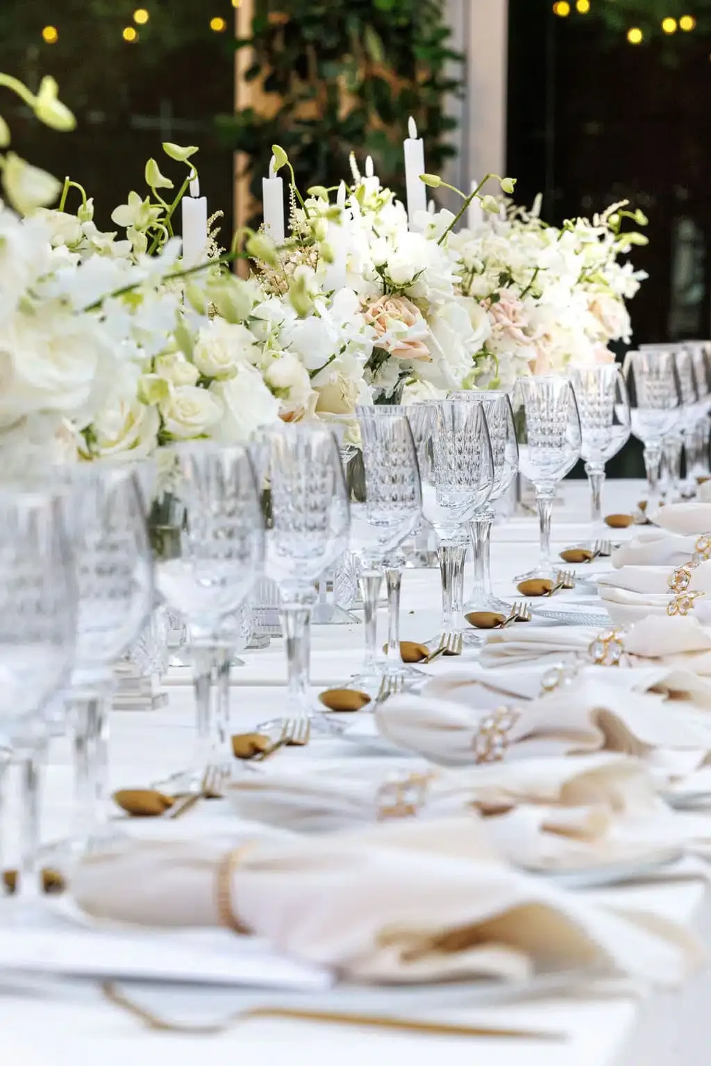 Elegant banquet table decorated with white floral arrangements and candles, set with crystal glasses, gold-toned cutlery, and white napkins for a formal event.