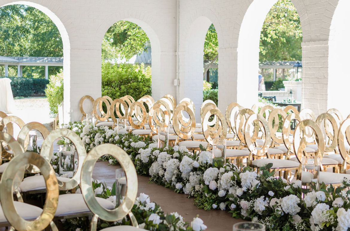 jksrentals Wedding ceremony setup with gold circular chairs and white floral arrangements inside a white brick venue with large arched windows overlooking greenery.