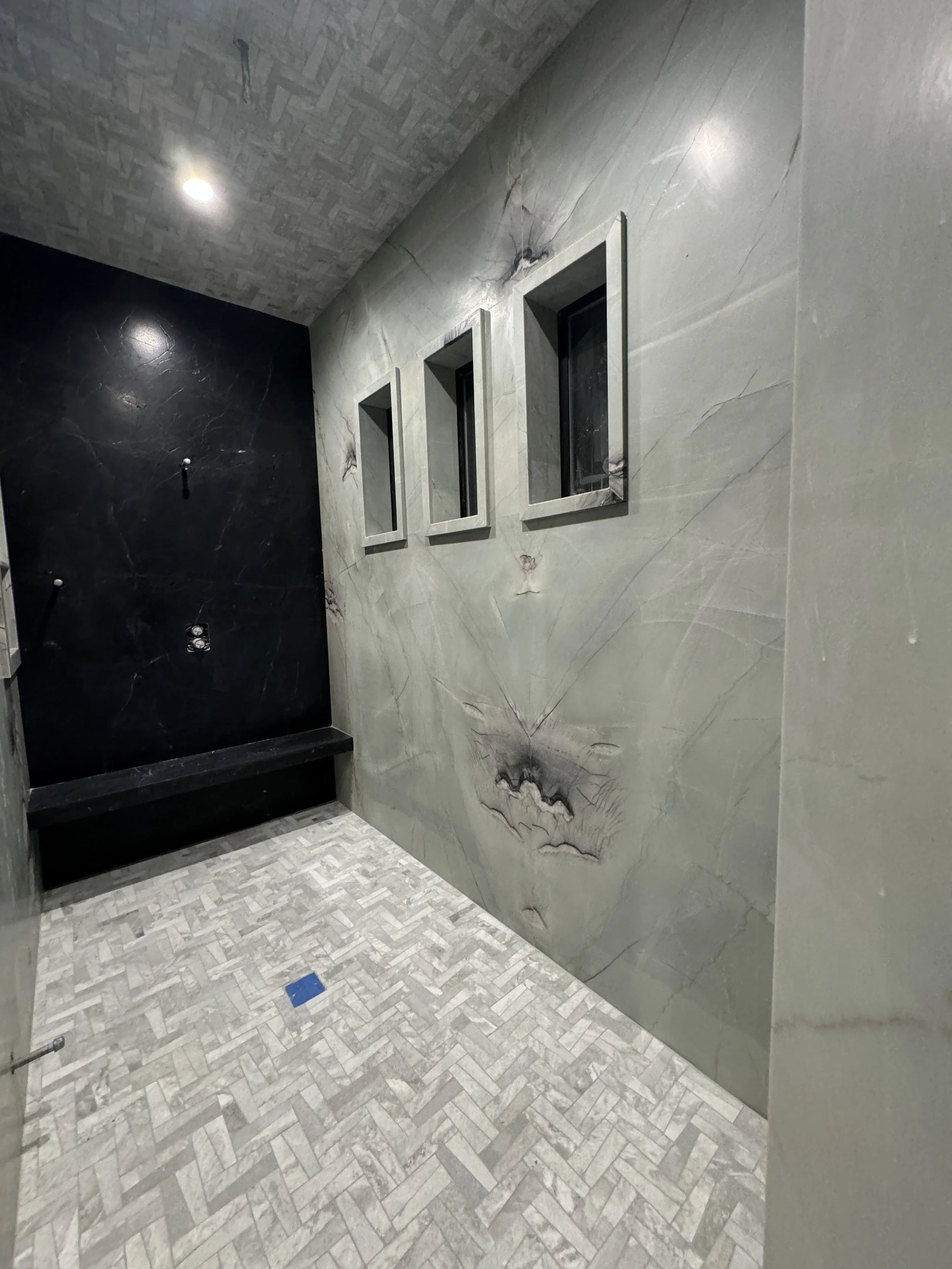 Empty shower area with black and grey marble walls, three narrow windows, and herringbone patterned tile floor.