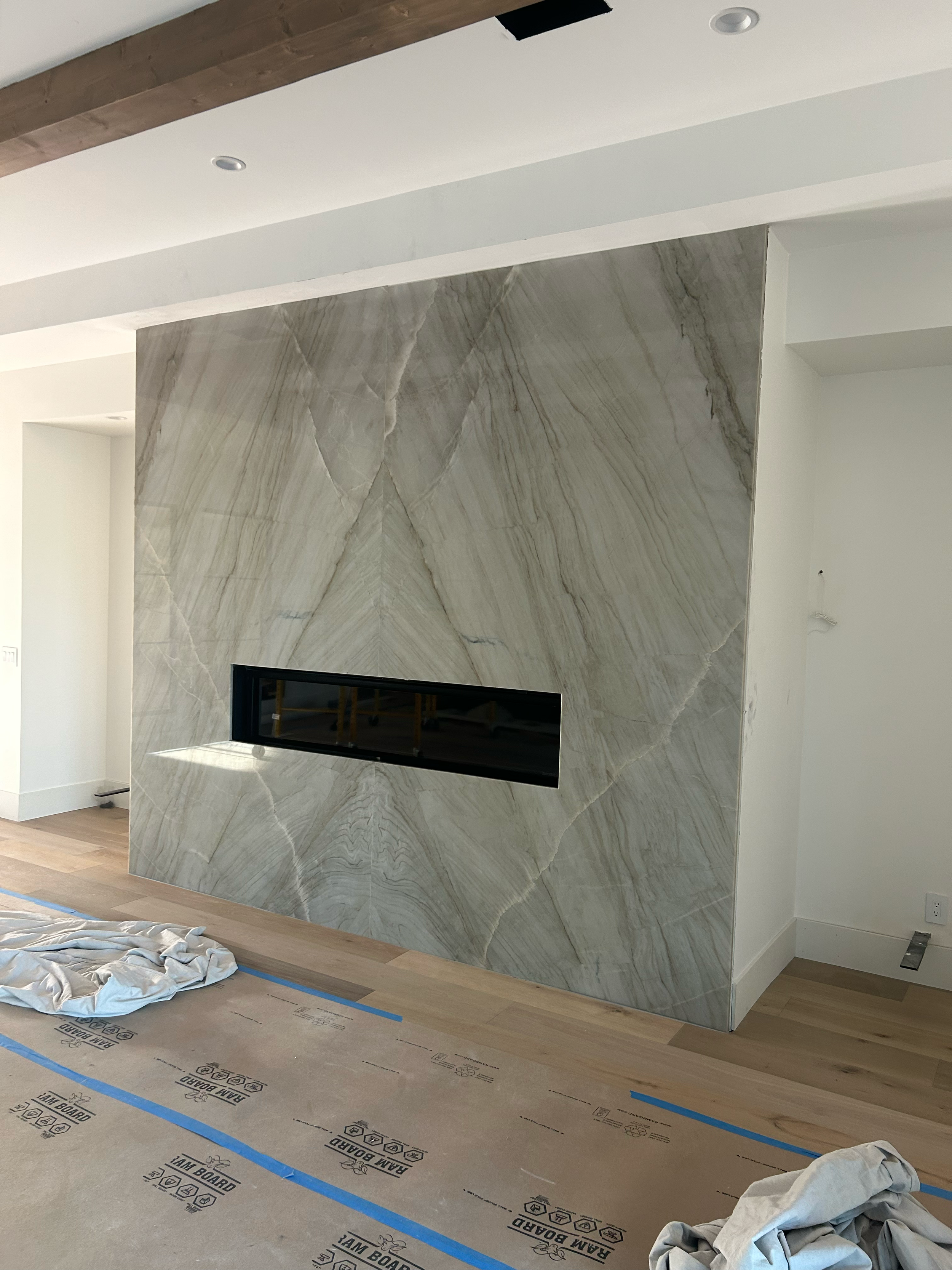A modern interior living space featuring a large marble fireplace with a sleek black insert, surrounded by white walls and a wooden ceiling beam, with protective covering on the floor indicating ongoing construction or finishing work.
