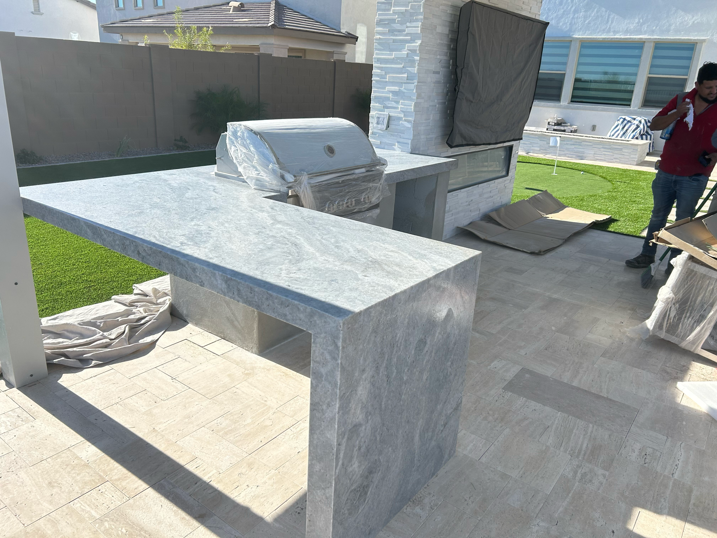 Outdoor kitchen area with a stone countertop, grill covered with plastic, and a man standing nearby holding a cloth in a backyard with a green lawn.