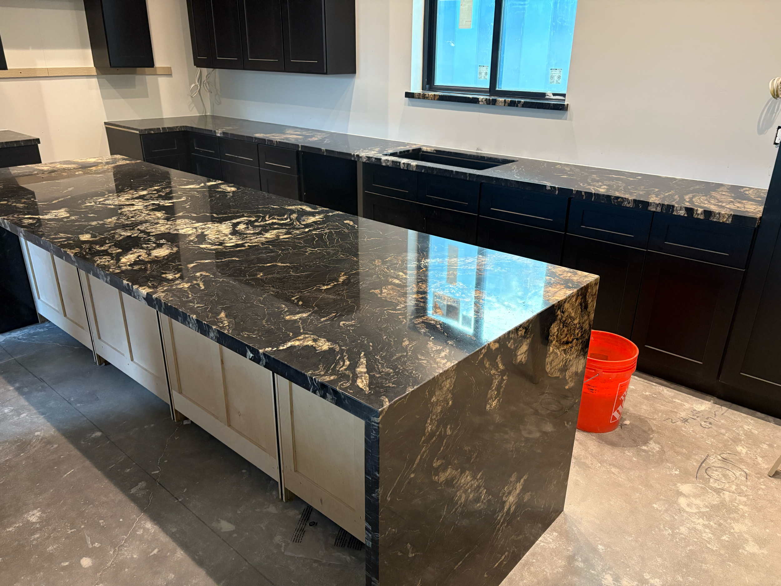 Kitchen with black and brown marble countertops, black upper cabinets, and beige lower cabinets, under a window with blue-tinted glass.