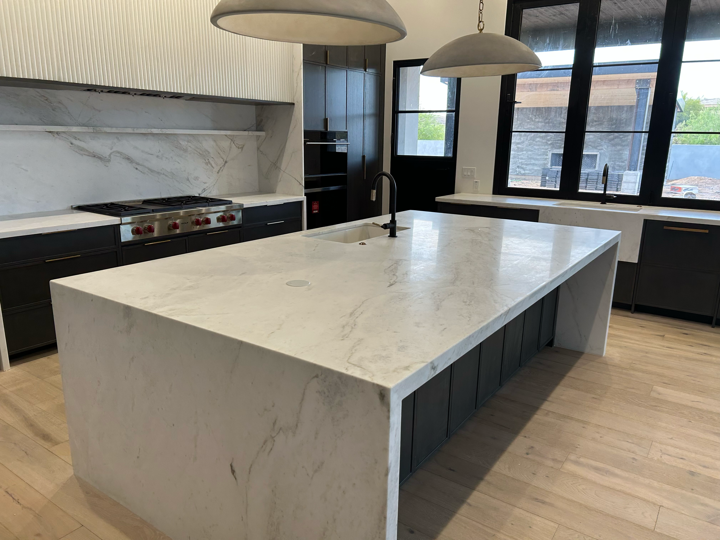 Modern kitchen with large white marble island, black cabinets, stainless steel stove with red knobs, black faucet, large windows, wood flooring, and pendant lights.