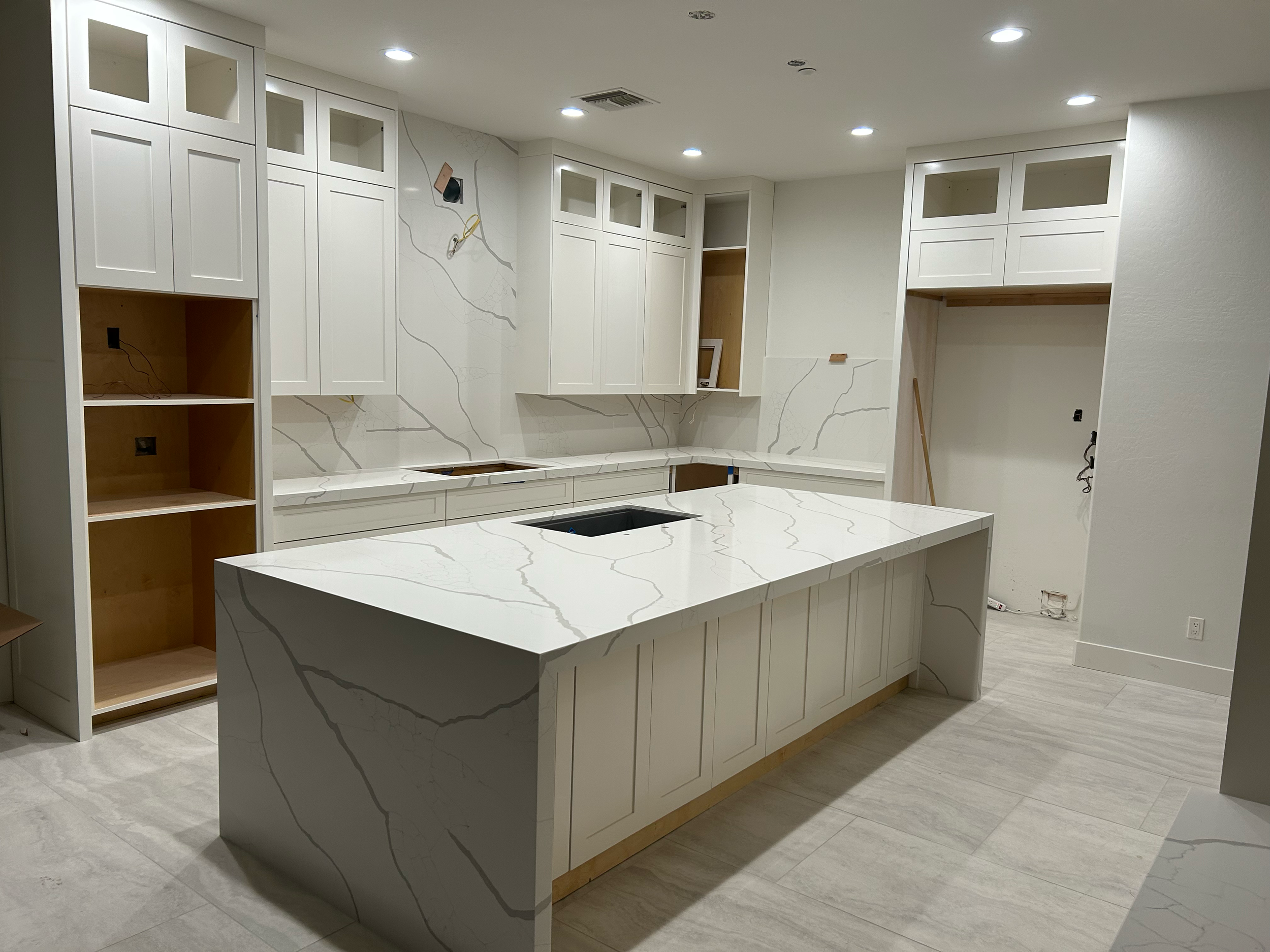 Modern kitchen under construction with white cabinets, marble countertops, and an island, with some appliances and wall spaces still unfinished.