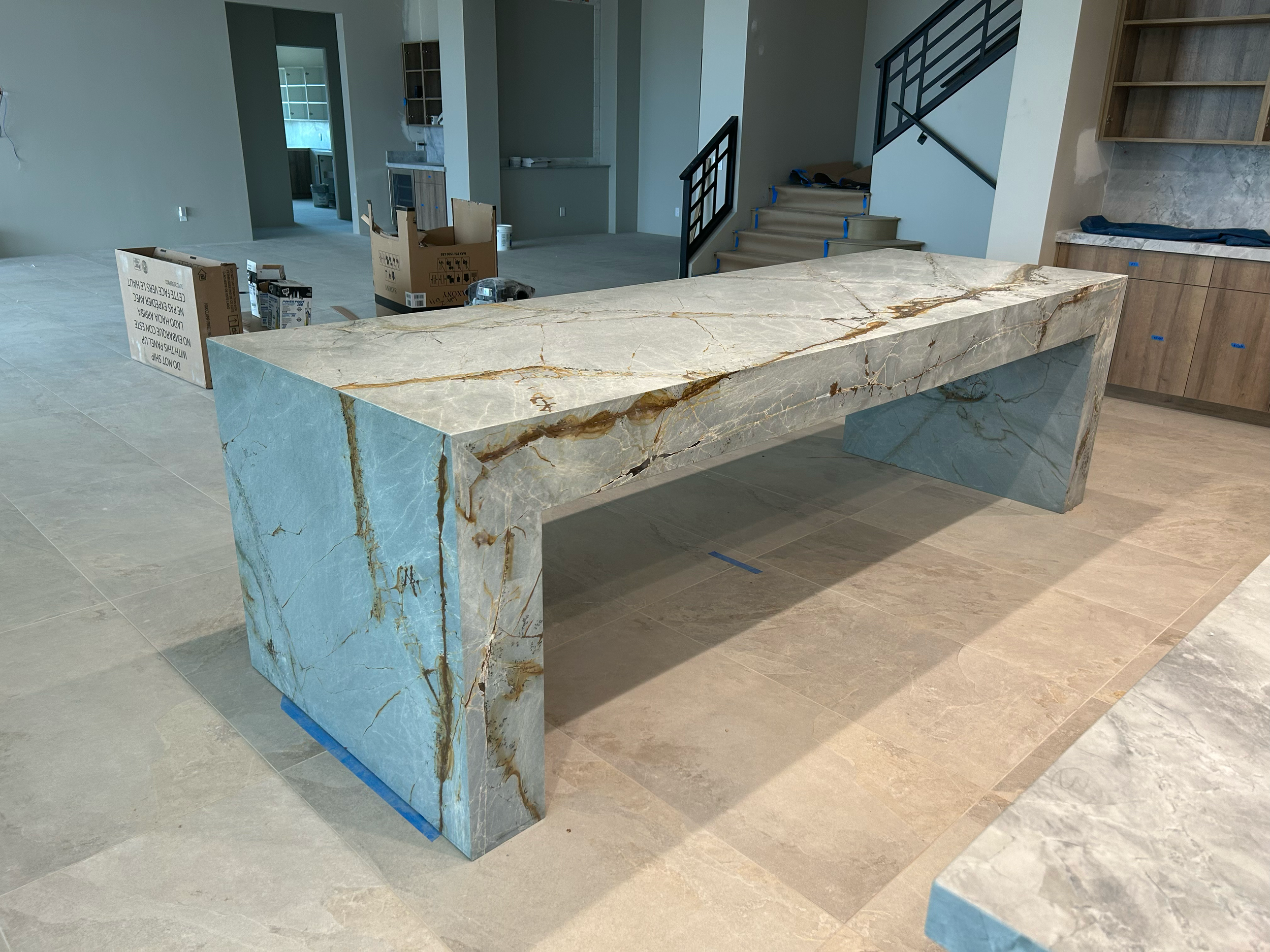 A marble kitchen island with a rectangular shape and a marble top, situated in a modern, under-construction kitchen. The island has a stone base with a matching marble countertop, and the surrounding area shows ongoing construction with boxes, tools, and partially installed cabinetry.