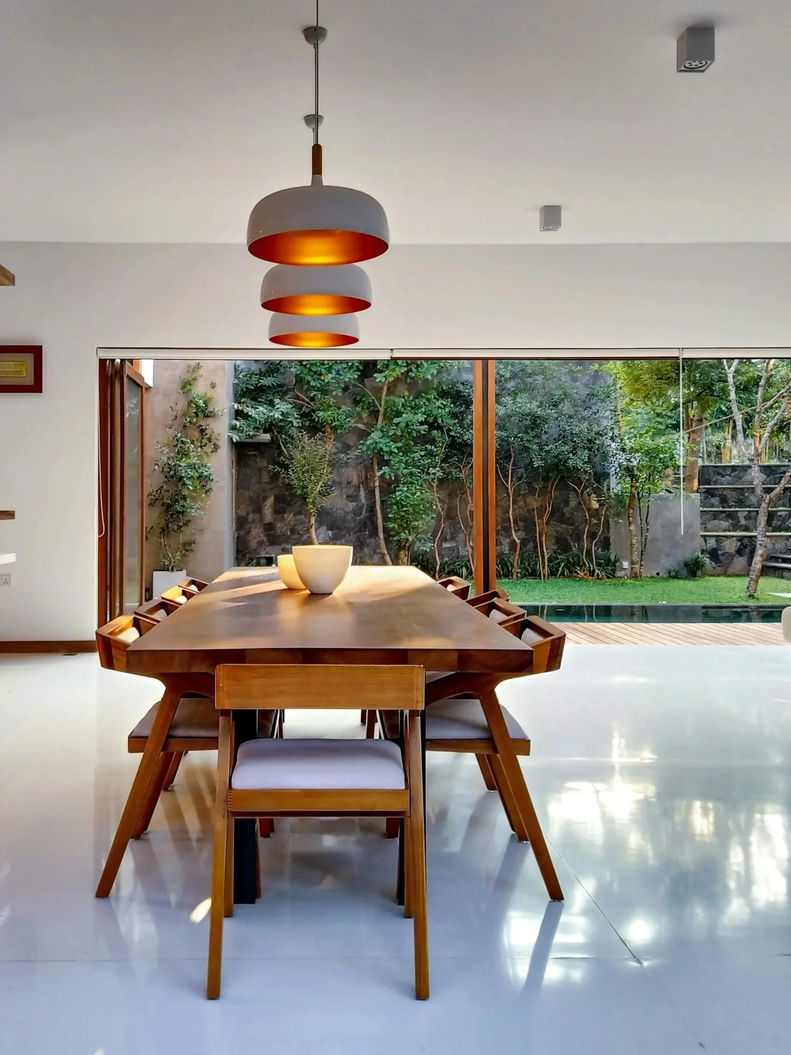 Modern dining room with a wooden table and chairs, hanging pendant lights, and large sliding glass doors opening to a lush green garden.
