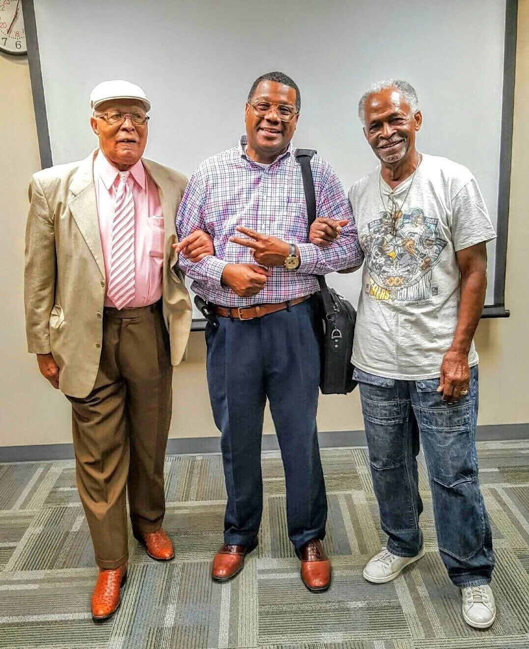 Three men standing together indoors in front of a whiteboard. The man on the left wears a beige suit with a pink shirt, striped tie, and a white hat. The man in the middle wears glasses, a checkered shirt, and jeans, carrying a black shoulder bag. The man on the right wears a gray graphic t-shirt and jeans. They are all smiling and holding hands.