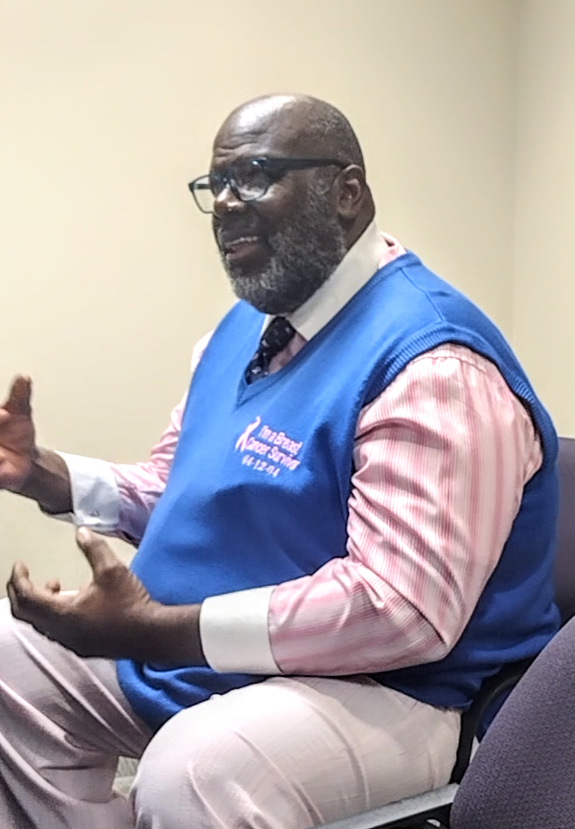 A man with glasses, a beard, and a bald head, wearing a blue vest over a pink striped shirt and beige pants, sitting and speaking animatedly in a room with plain yellow walls.