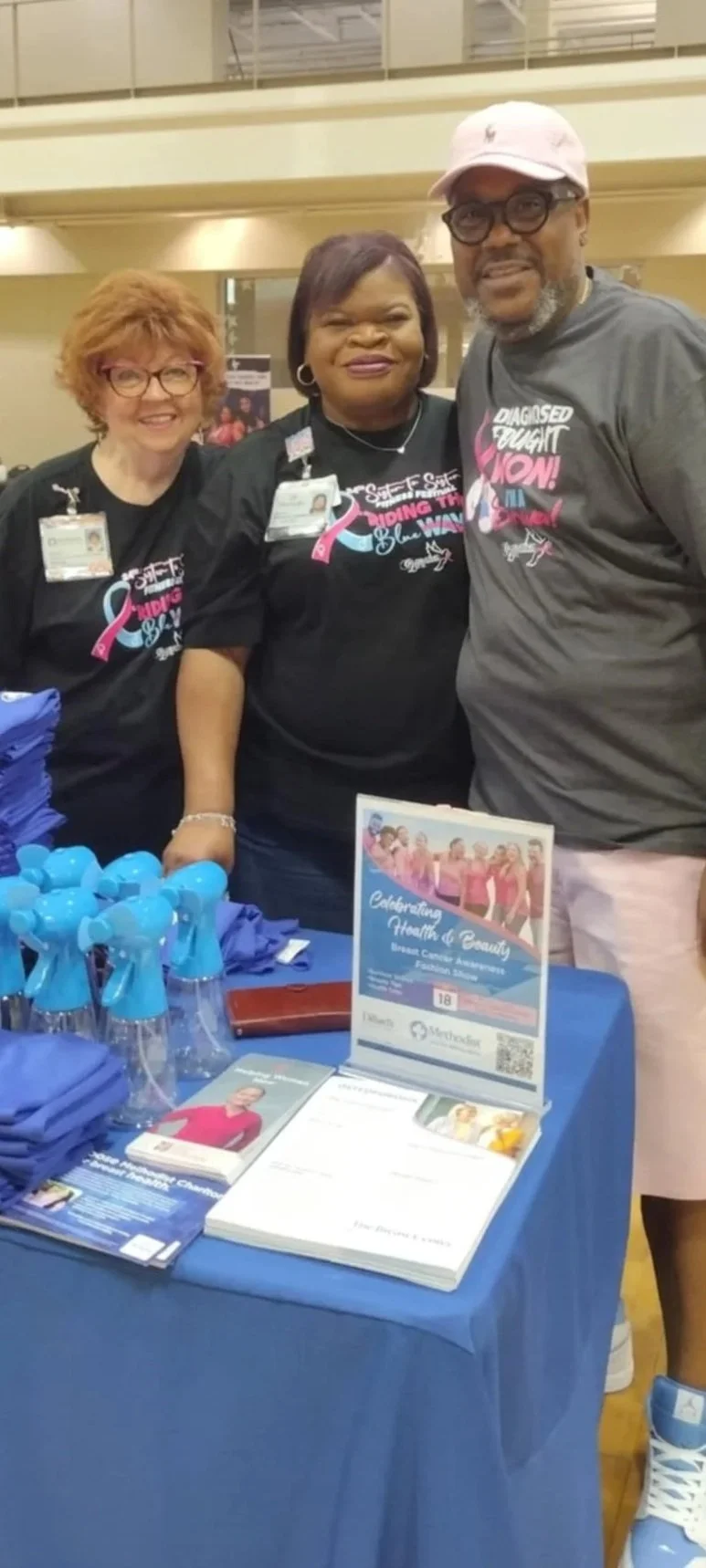 Three people at a health awareness event, standing behind a table with informational materials, wearing black T-shirts with pink and white text supporting breast health awareness.