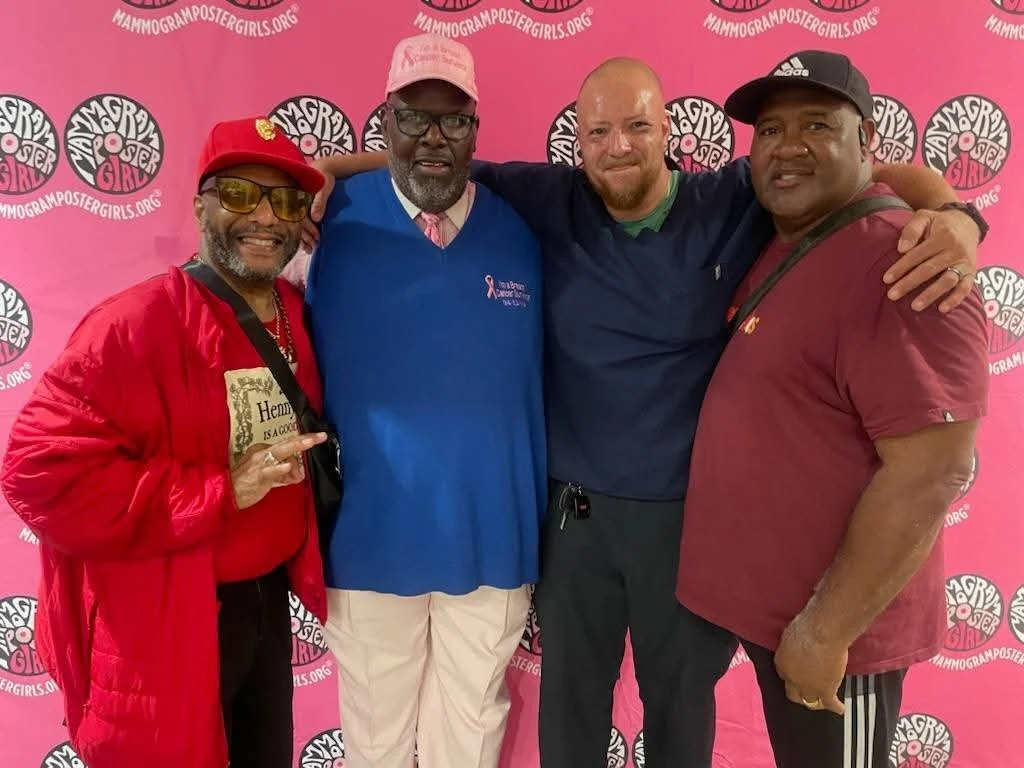 Four men standing together at a breast cancer awareness event against a pink background with the 'Mammogram Poster Girls' logo. They are smiling and have their arms around each other, dressed casually, with one man wearing a red jacket and hat, anoth
