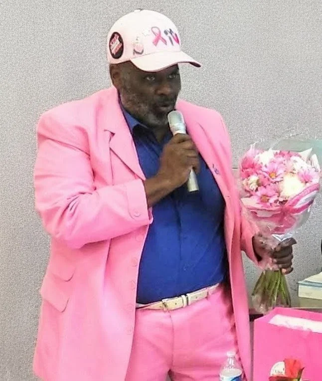 Man in pink suit, wearing a pink hat with a breast cancer awareness ribbon, holding a microphone and a bouquet of pink flowers.