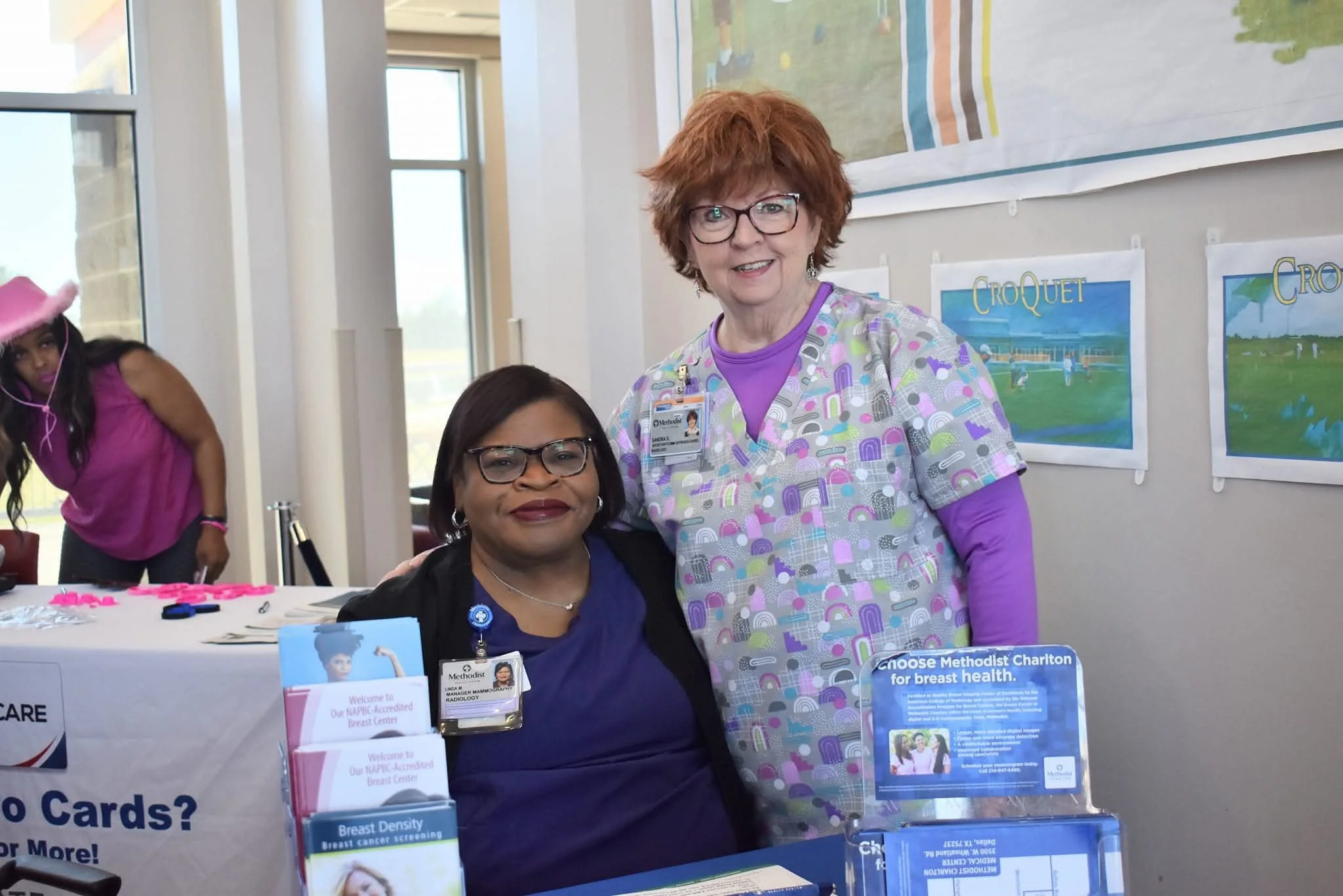Two women at a health fair booth, one seated and one standing, with pamphlets and informational displays about breast health and screening.