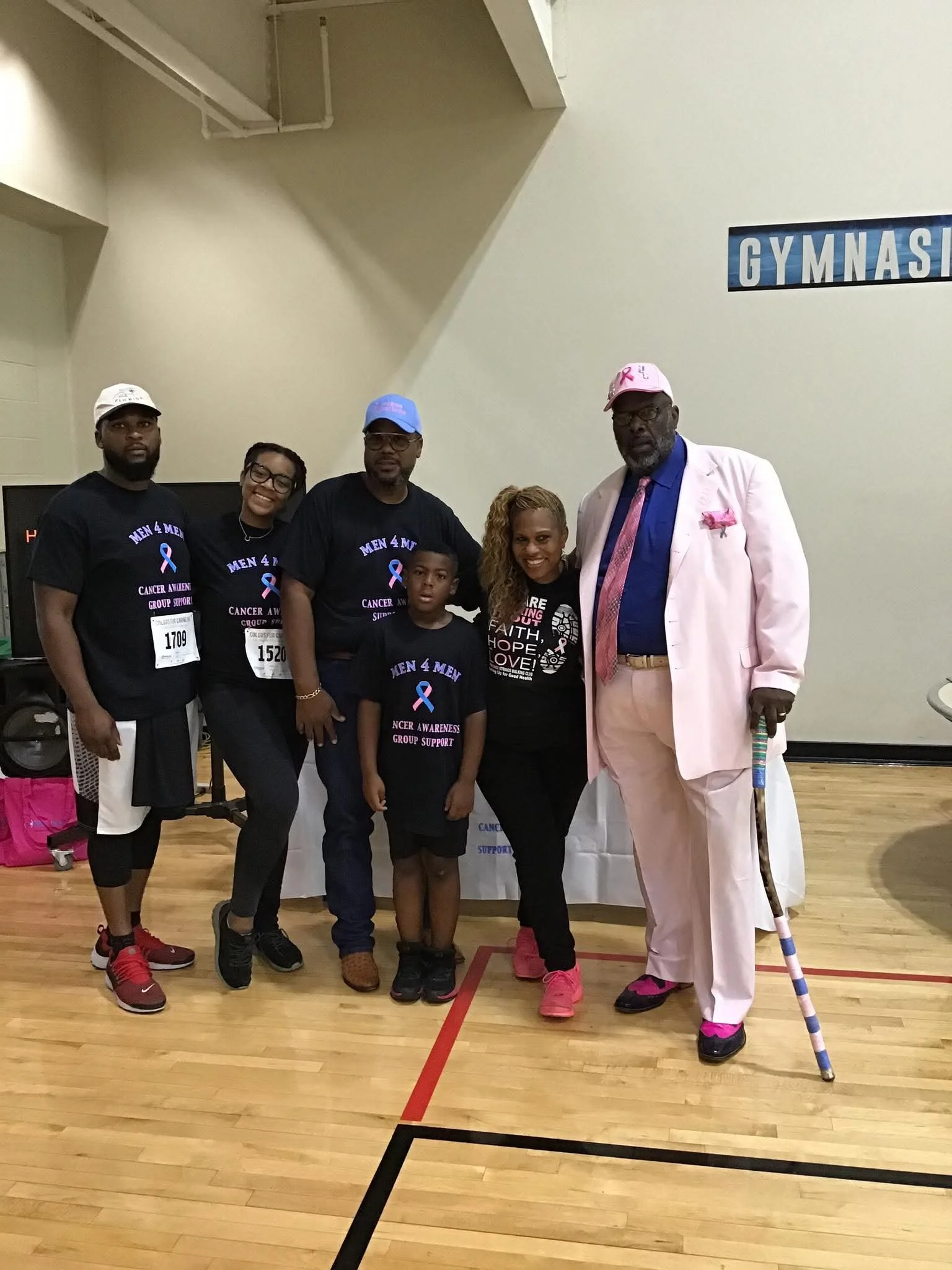 Group of six people standing in a gymnasium, wearing awareness shirts for cancer, with some wearing caps, and an elderly man in a pink suit with a cane.