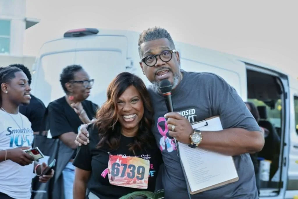 A group of people at a charity event, with two individuals in the foreground smiling and posing for the camera, one holding a microphone, the other wearing a race bib number 639.