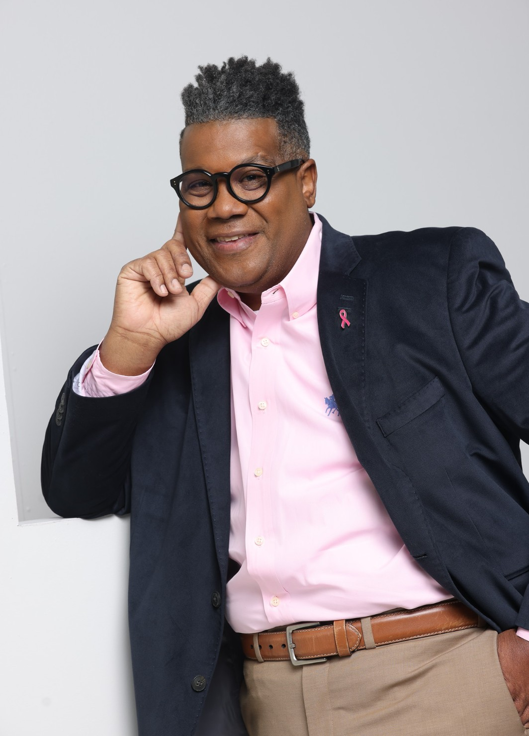 A man with glasses and short curly hair, wearing a pink button-up shirt, dark blazer, and beige pants, smiling and leaning against a white wall with his hand near his face.