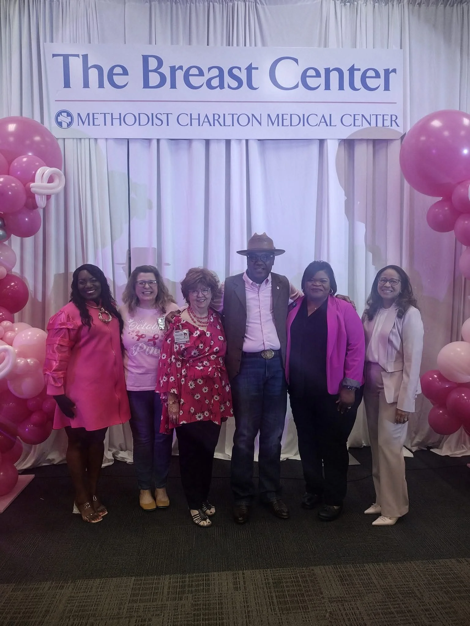 Group of six women and one man standing together in front of a sign that reads 'The Breast Center, Methodist Charlton Medical Center.' The background is decorated with pink and white balloons and curtains, indicating a breast cancer awareness event o