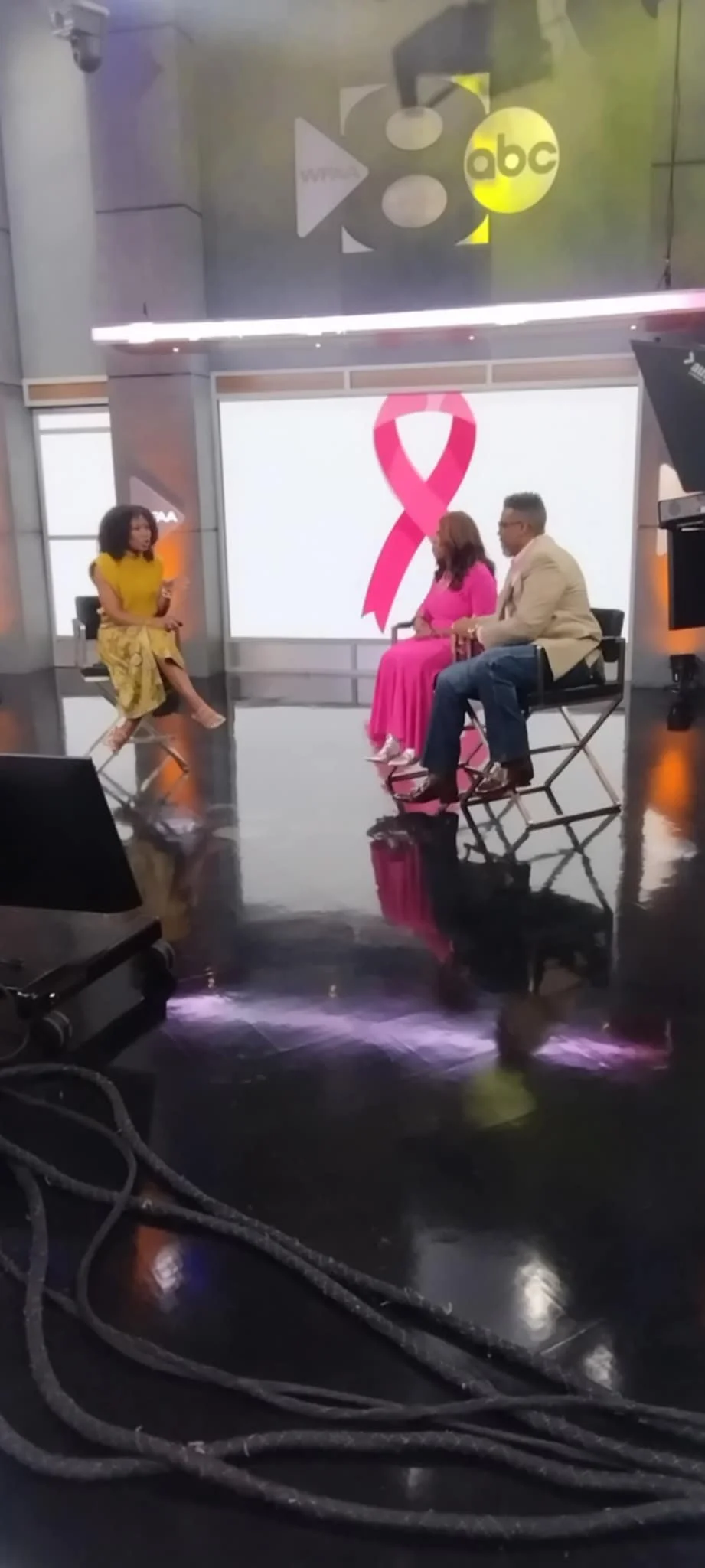Television studio with three people in discussion, large breast cancer awareness pink ribbon on screen, and NBC logo overhead.