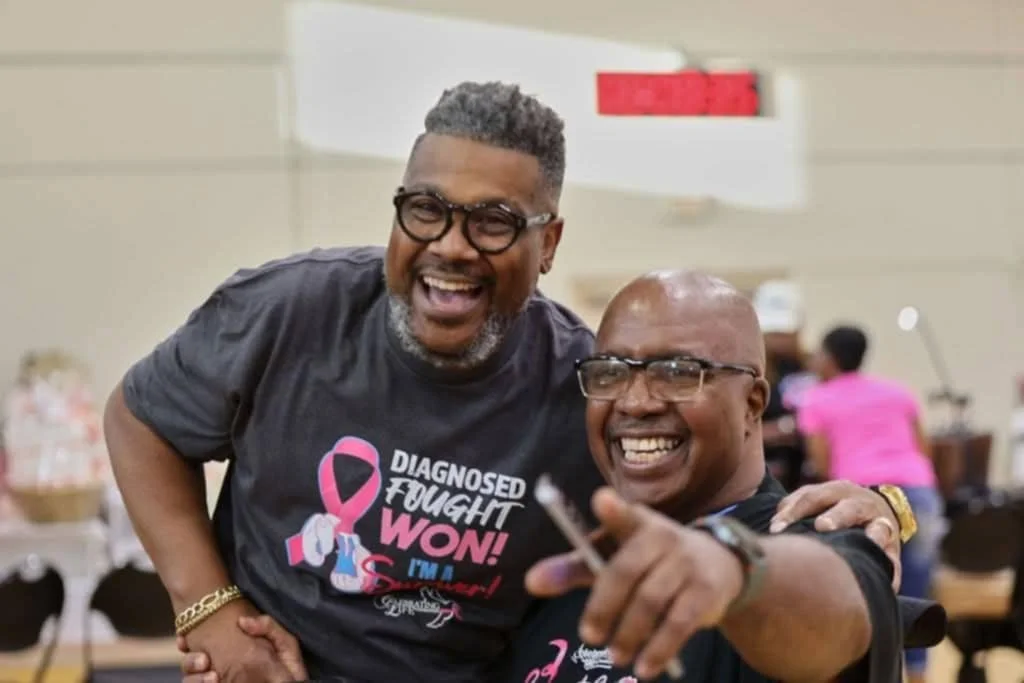 Two men smiling and posing for a photo at an indoor event. One man is wearing a black t-shirt with pink text about breast cancer awareness and the other is wearing a similar shirt, pointing at the camera.