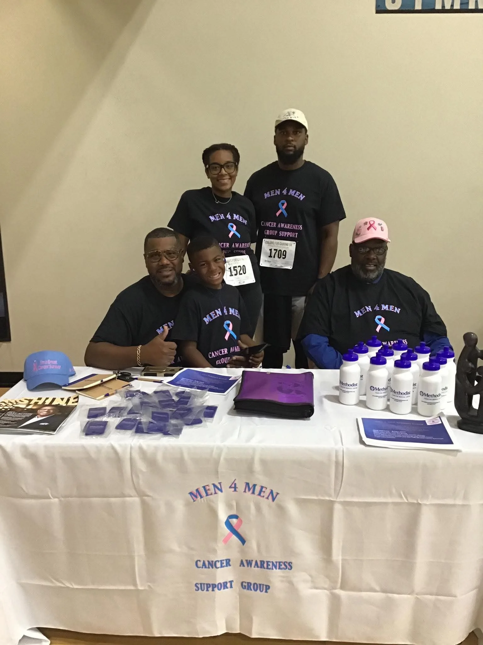 Group of five people wearing black 'Men 4 Men' cancer awareness T-shirts, sitting and standing behind a table with informational materials and bottles of hand sanitizer, at a cancer awareness support event.