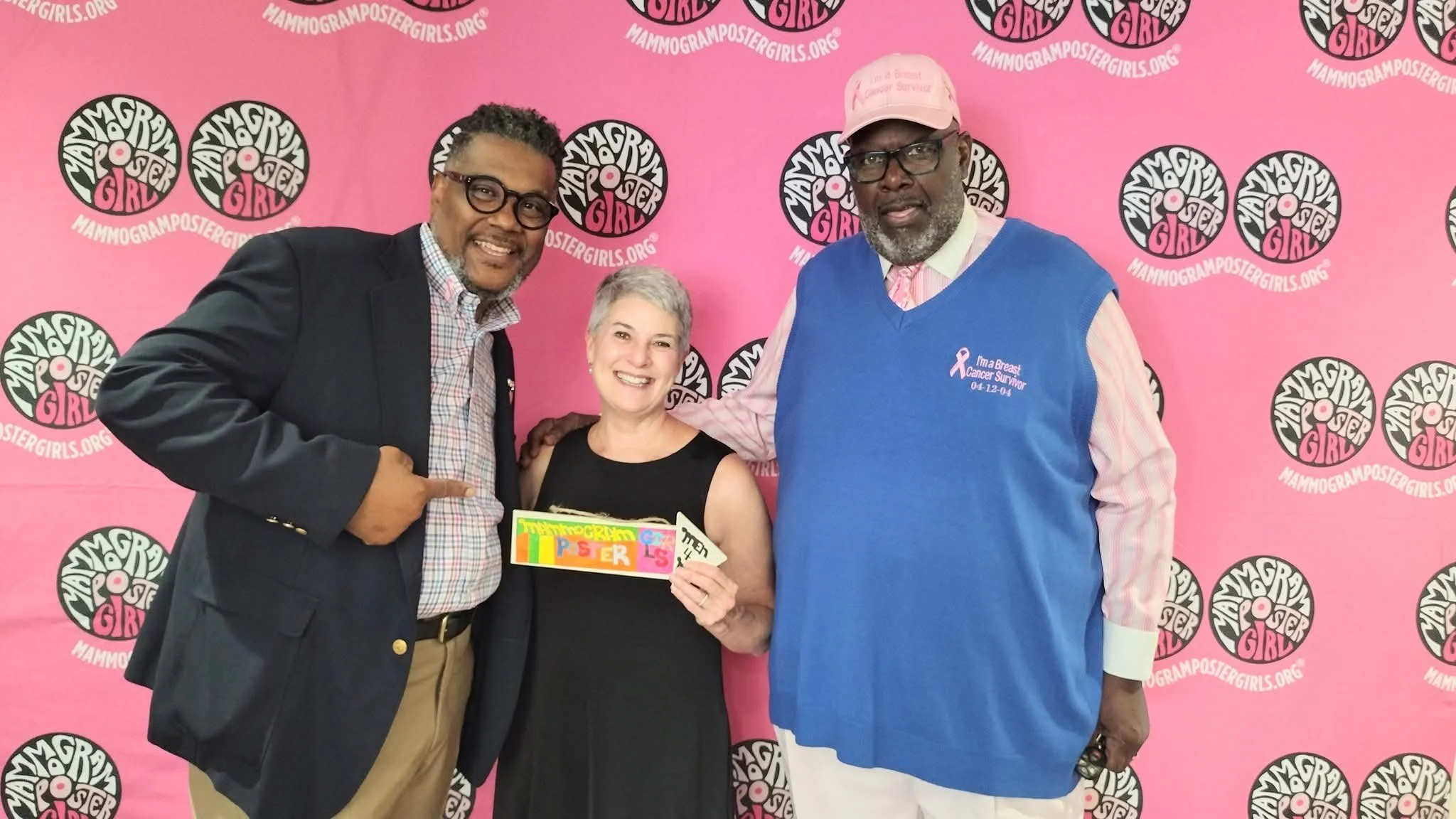 Three people standing together in front of a pink background with black and white logos that read "Mammogram Poster Girl," smiling and posing for the photo. The woman in the middle holds a colorful sign.