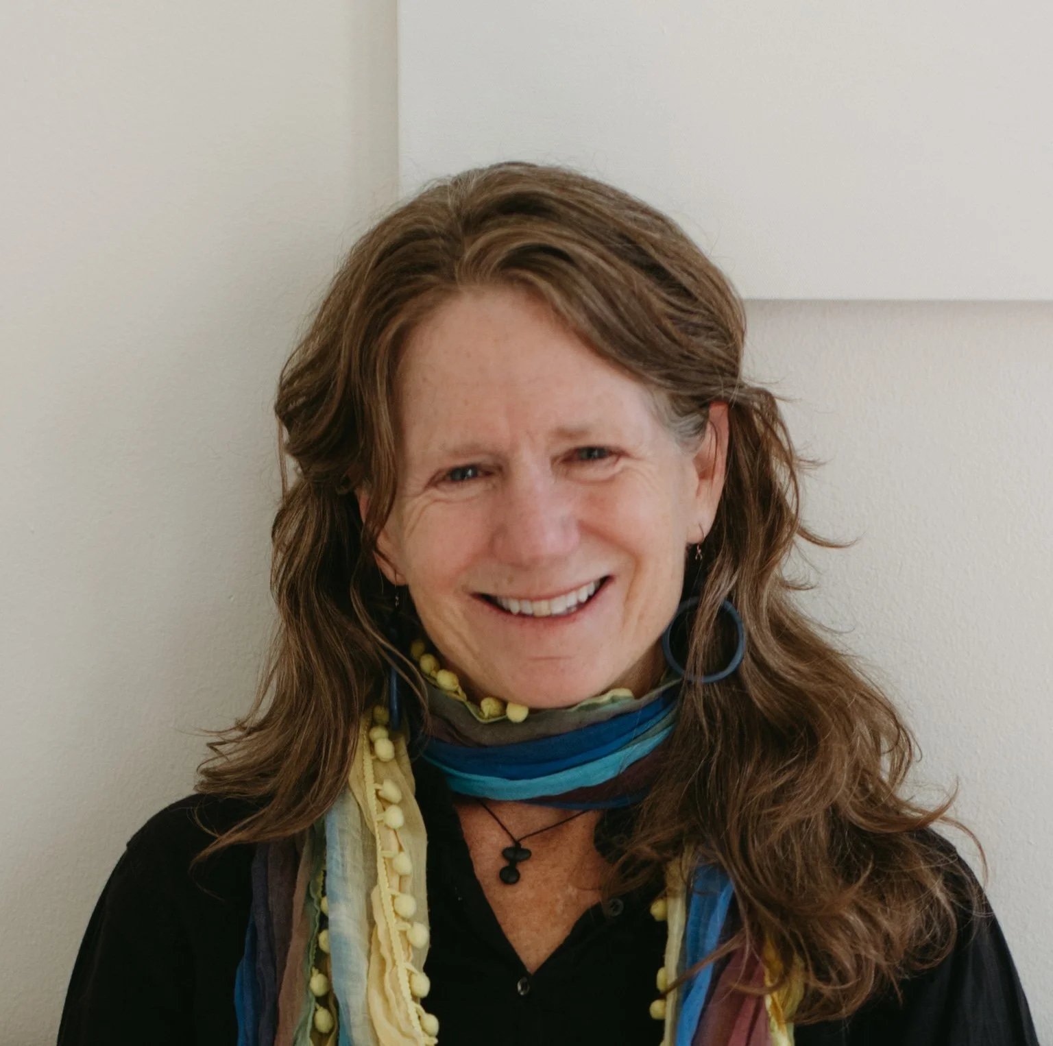 A woman with long, wavy brown hair, smiling, wearing a multicolored scarf and jewelry, against a plain light-colored wall.
