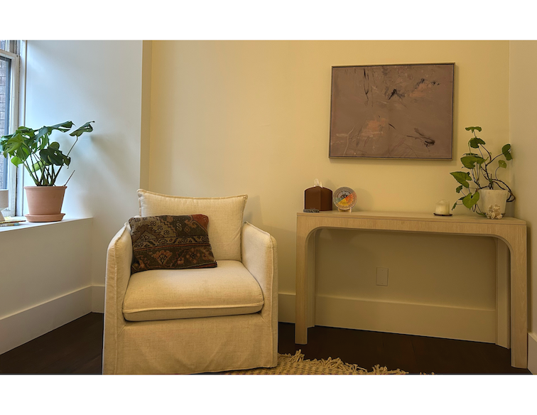 Cozy corner of a living room with a white armchair, decorative pillow, potted plant on the windowsill, beige wall, and a light-colored console table with small decorative items and a large abstract painting.