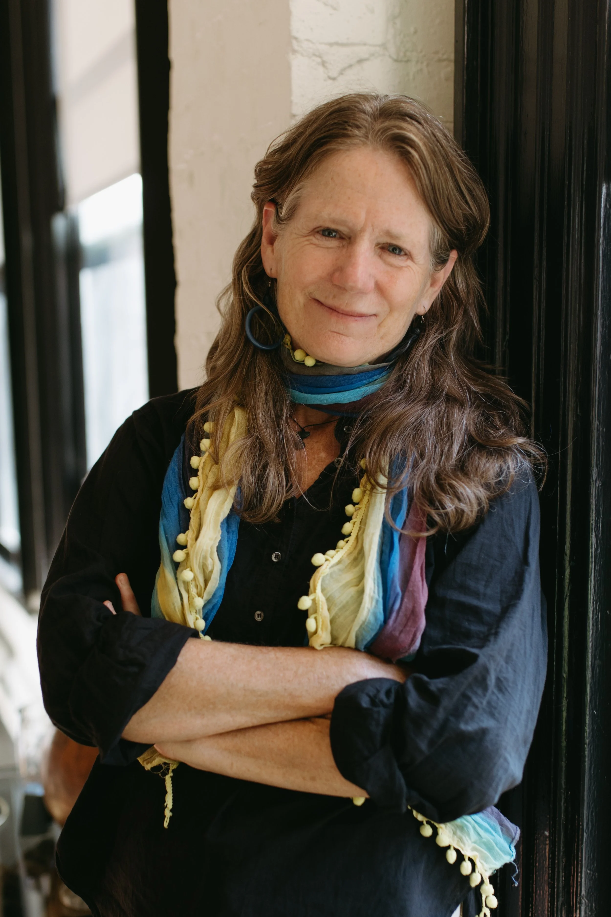A woman with long, wavy hair wearing a black shirt and colorful scarf, standing with crossed arms, smiling slightly, in front of a black door and light-colored wall.