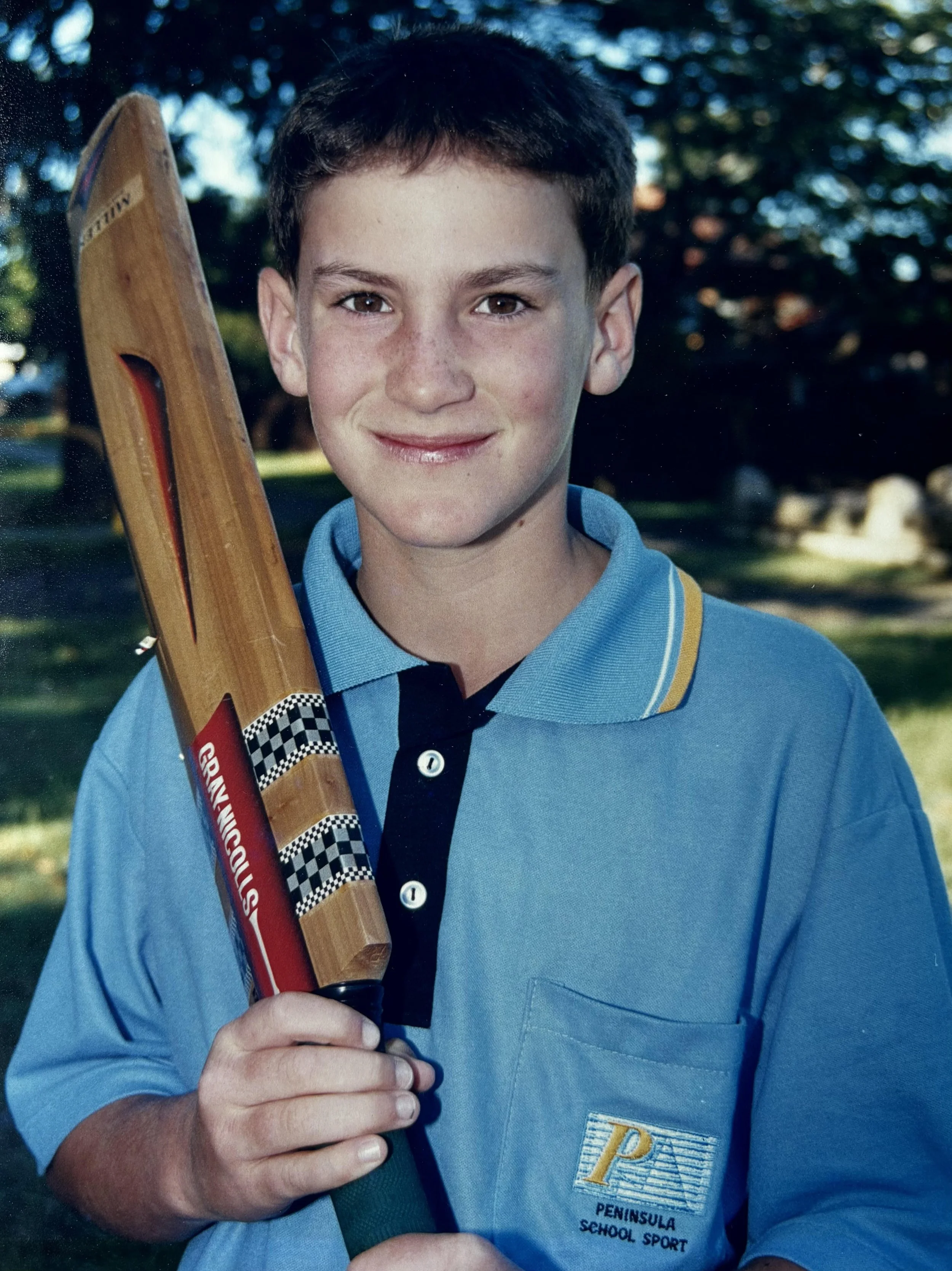 A young boy with short dark hair, wearing a blue polo shirt with a Peninsula School Sport logo, holding a wooden cricket bat with a red and checkered sticker, standing outdoors with trees in the background.