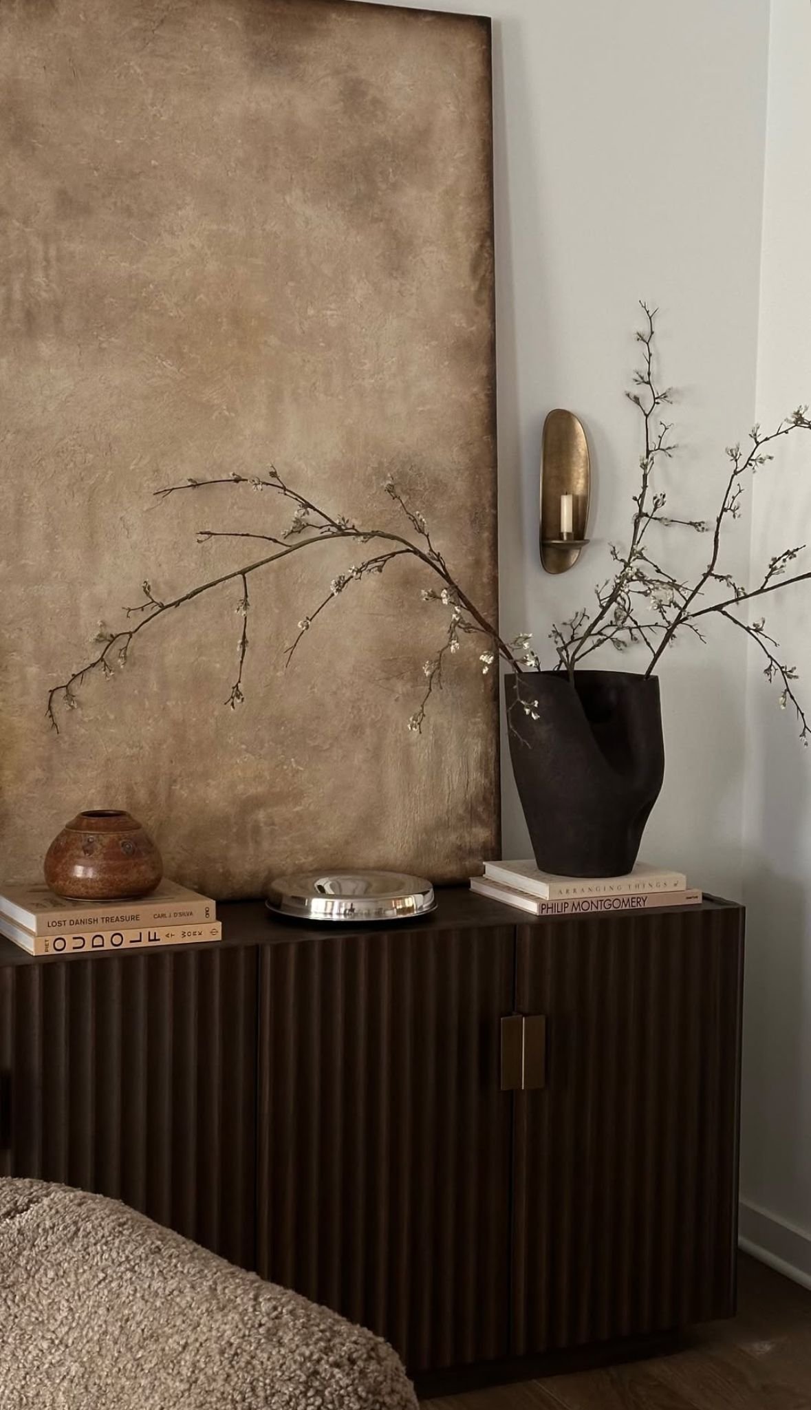 A beige textured wall with a black cabinet holding decorative items including a large black vase with branch-like plants, a small clay pot, a stack of books, and a silver tray. A gold wall sconce with a white candle is mounted on the wall next to the vase.