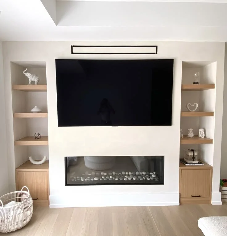 Living room entertainment wall with a mounted flat-screen TV above a modern fireplace with white rocks inside. Shelves on both sides holding decorative items such as art pieces, a sculpture, and books. A woven basket is on the floor to the left.