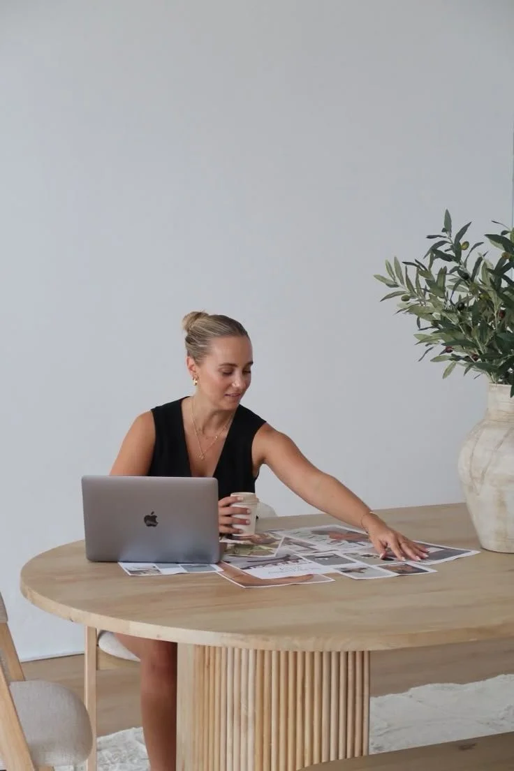 Woman sitting at a wooden table, reaching for papers with a laptop, holding a coffee mug, in a bright, minimalist room with a large potted plant.
