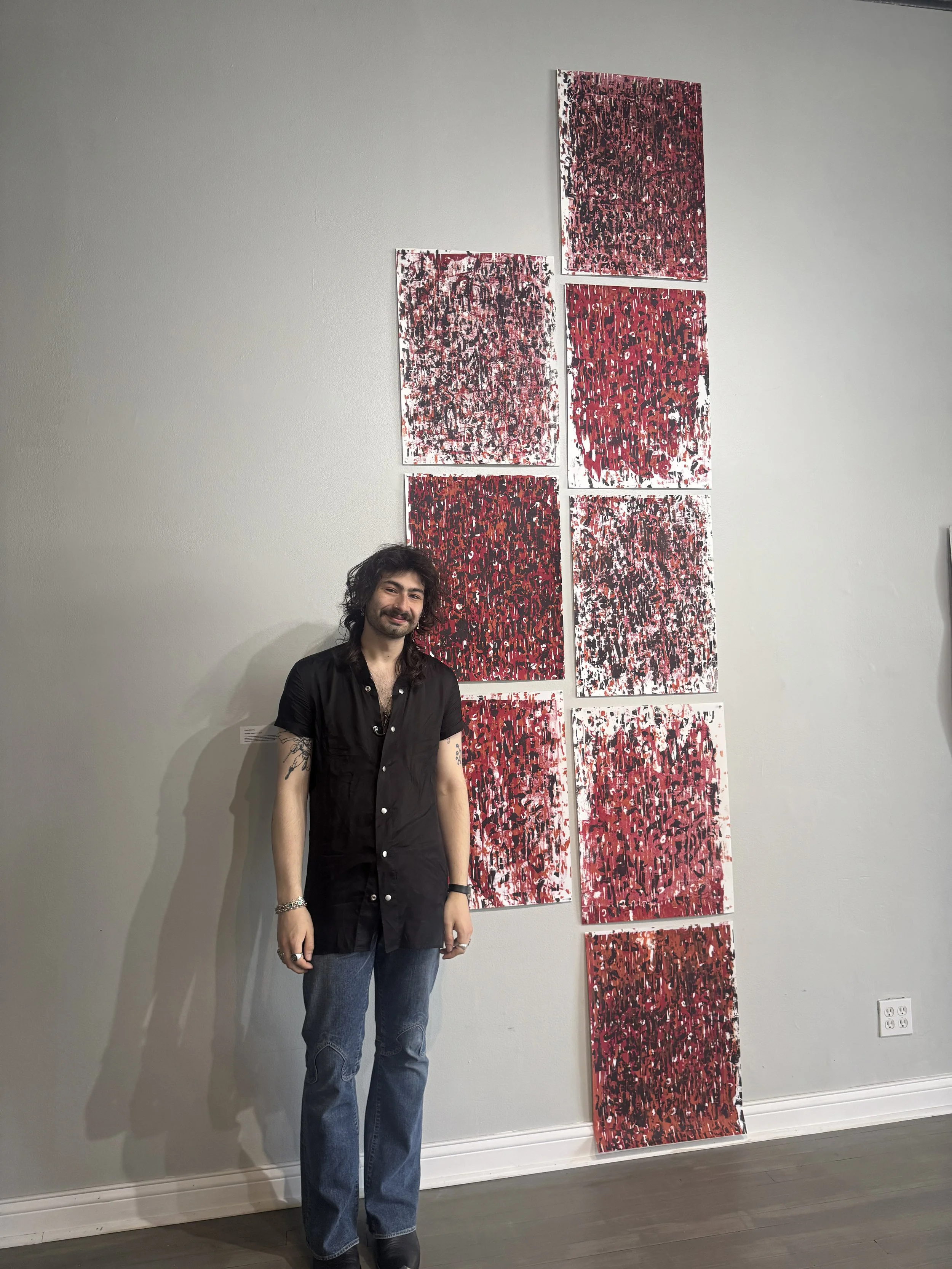 Man with long dark hair and beard standing next to an art display on a white wall, consisting of seven abstract paintings with red, black, and white color schemes.