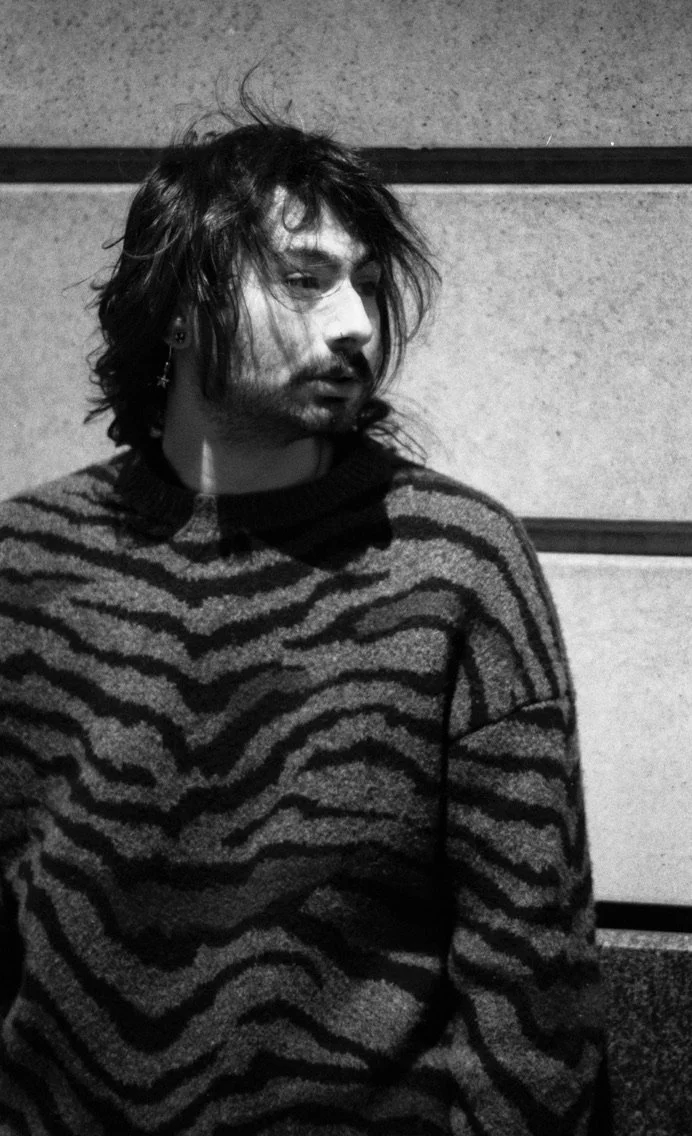 Black and white photo of a young man with shaggy hair, beard, and earrings, wearing a striped sweater, sitting against a textured wall.