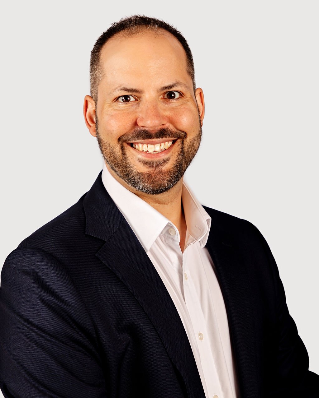 Professional headshot of a man with short hair, beard, wearing a white shirt and black blazer, smiling against a plain white background.