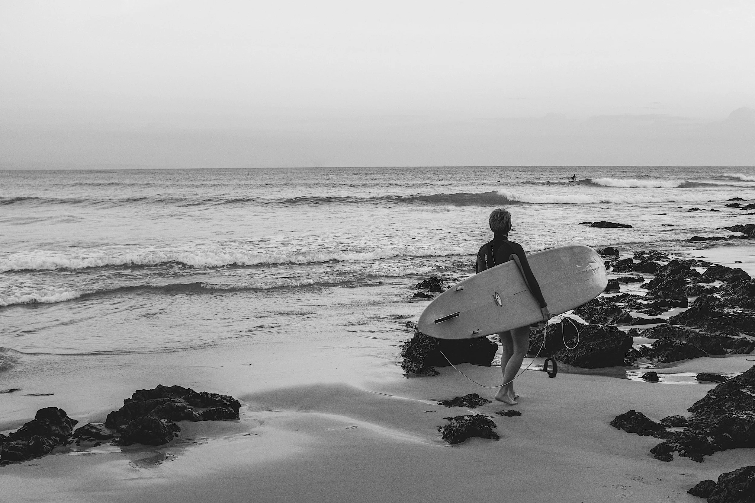 Black and white photo of a young person with a surf board looking out towards the surf and watching other surfers Byron Bay Australia