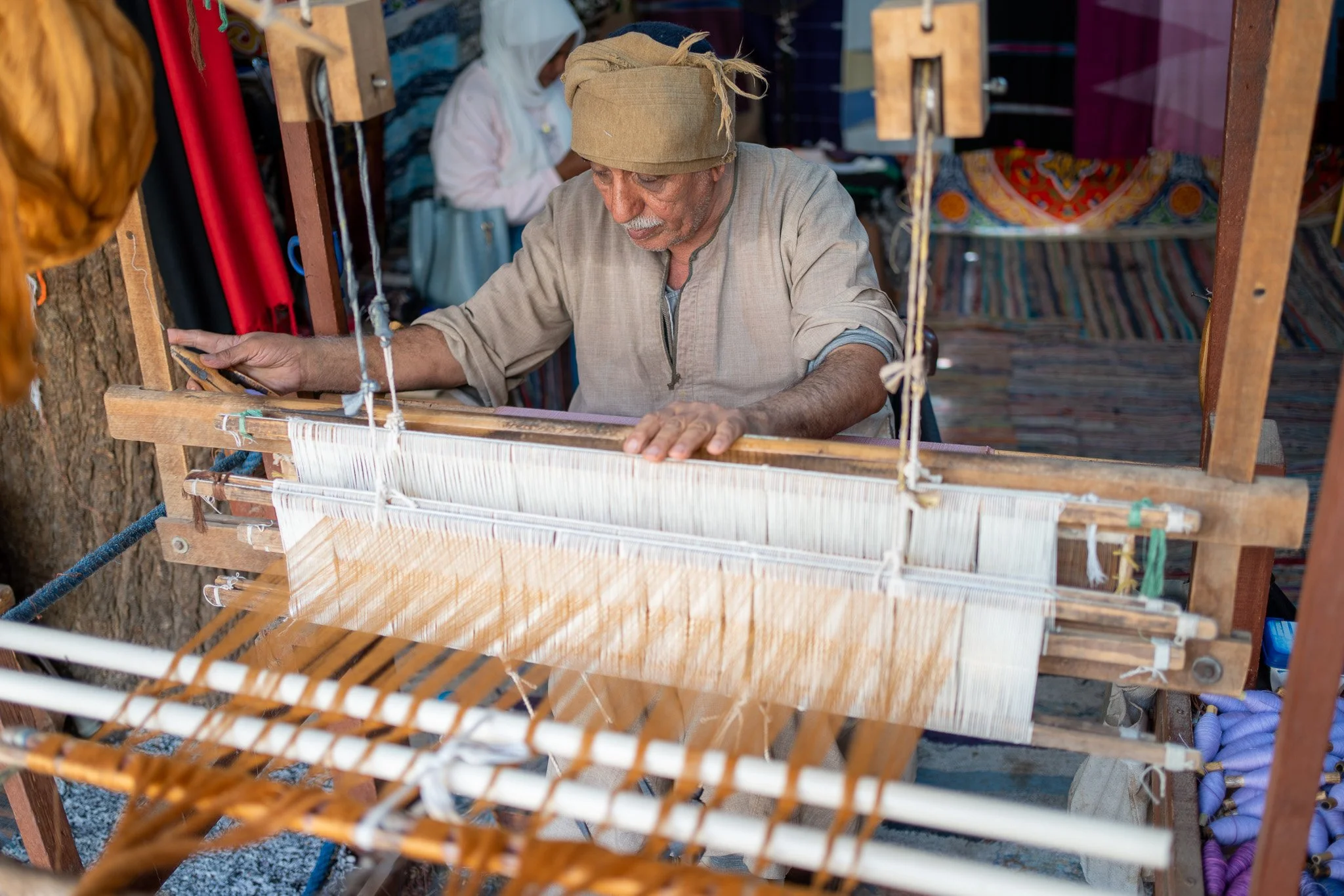 An older man weaving on a traditional wooden loom at an outdoor market.