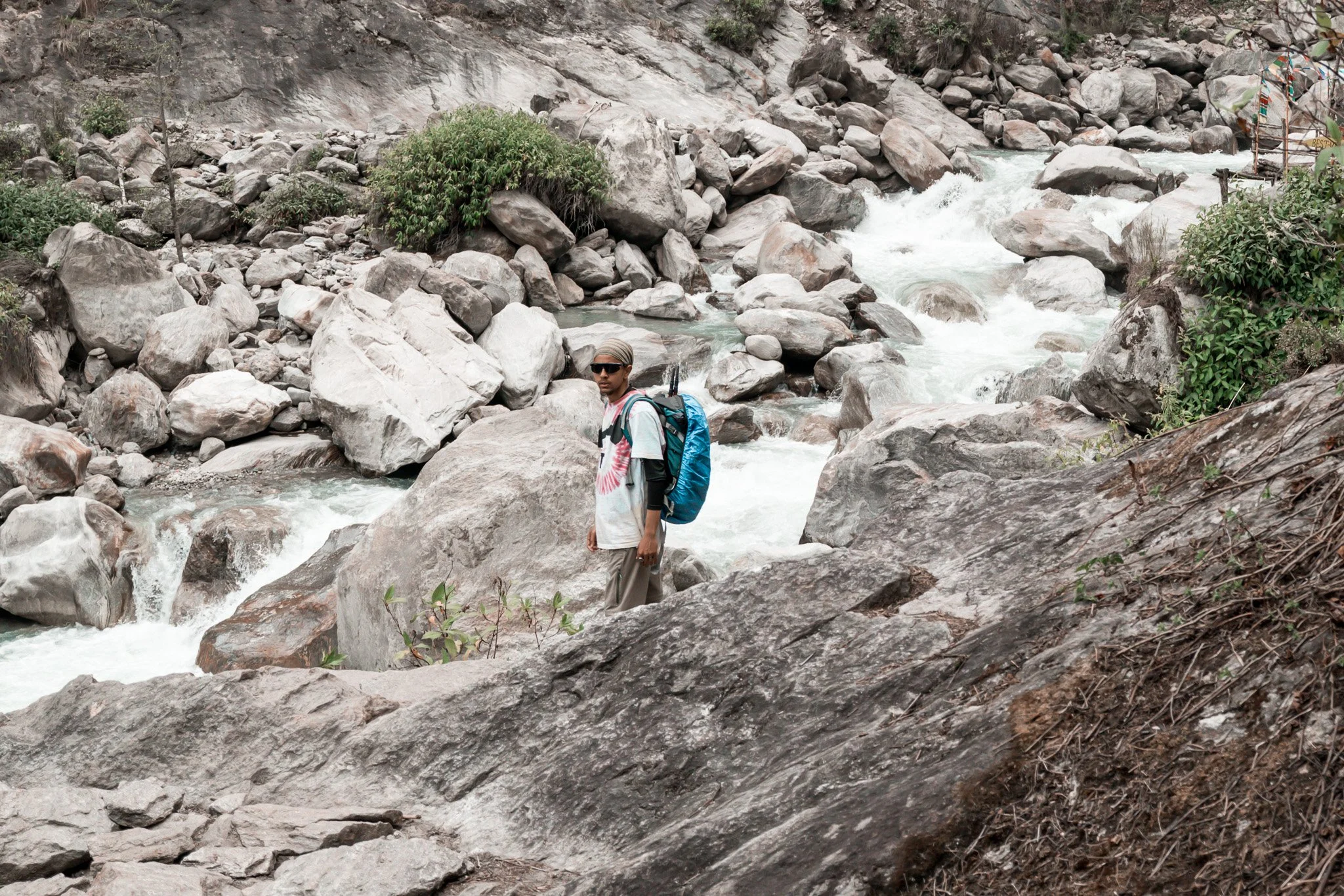 A man with a backpack standing on rocks next to a rushing mountain stream, rocky terrain, and some greenery.