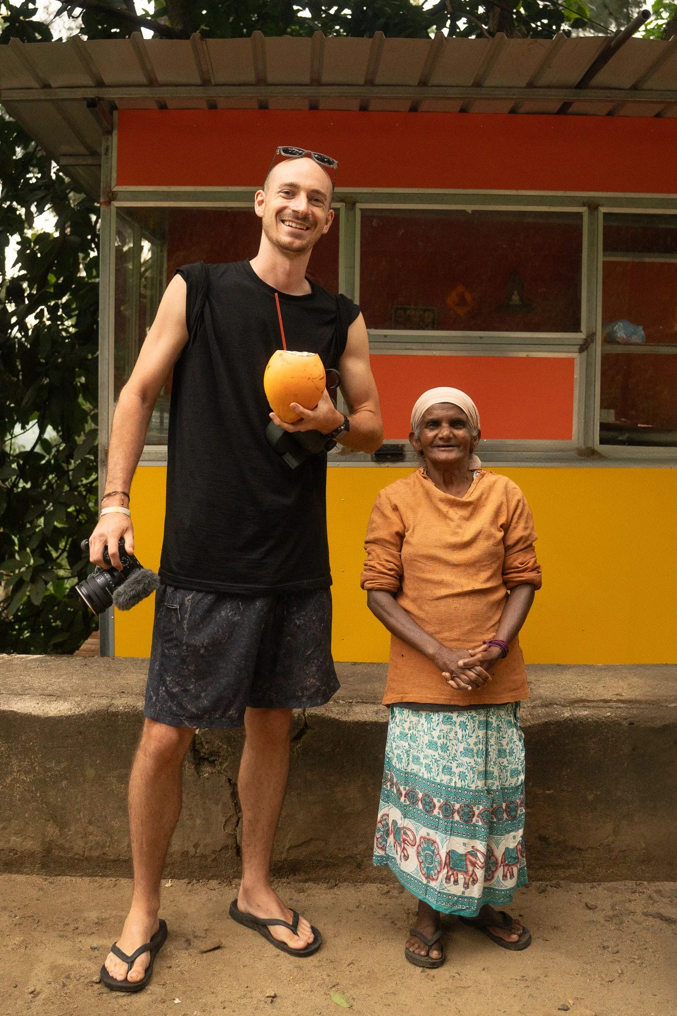 A young man with sunglasses on his head, holding a coconut with a straw, standing next to an elderly woman in traditional Indian attire, both smiling outdoors in front of a small food or snack stall.